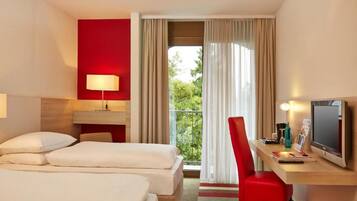 A hotel room with two beds, a desk with a computer, a chair, and a view of greenery through the window.