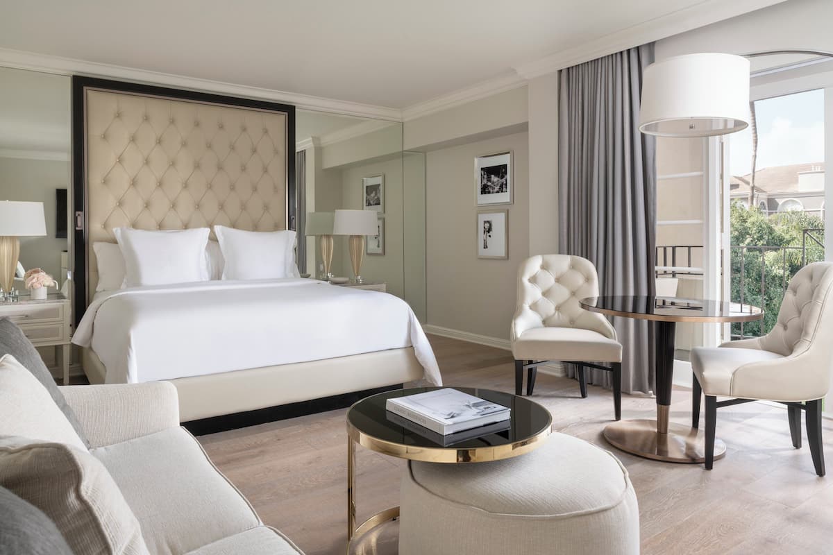 A modern hotel room with a large bed, a round coffee table, two armchairs, and a view of the outdoors.