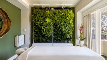 Una habitación de hotel moderna con una cama grande, un jardín vertical en la pared, una mesita con un florero y una ventana con cortinas.