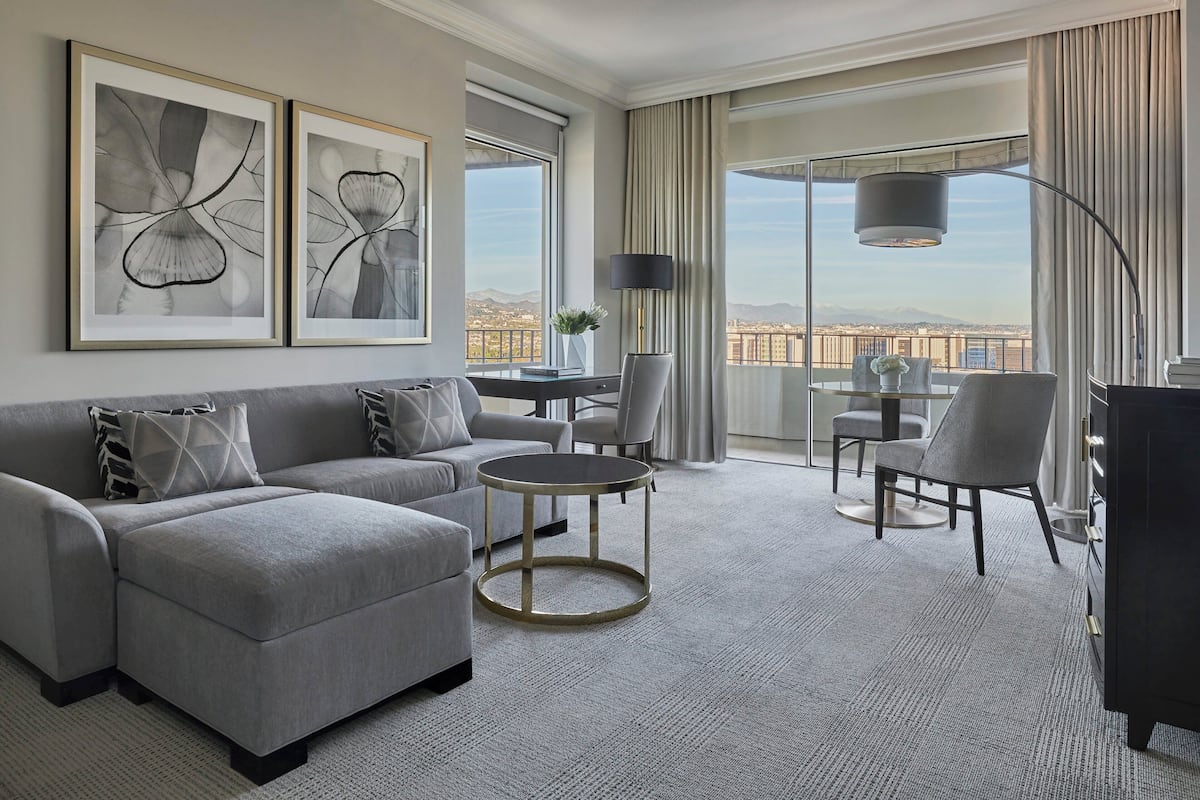 A modern hotel room with a grey sofa, a round coffee table, and a view of a cityscape through large windows.