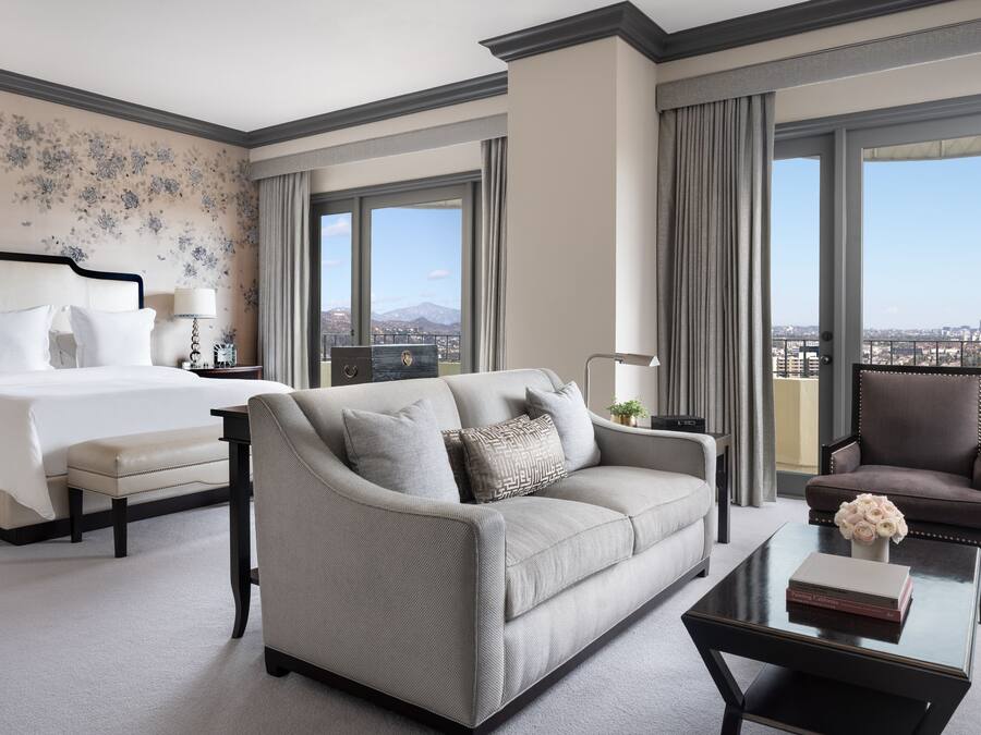 Quarto de hotel com cama, sofá, poltrona, mesa de centro e vista para a cidade através de janelas amplas.