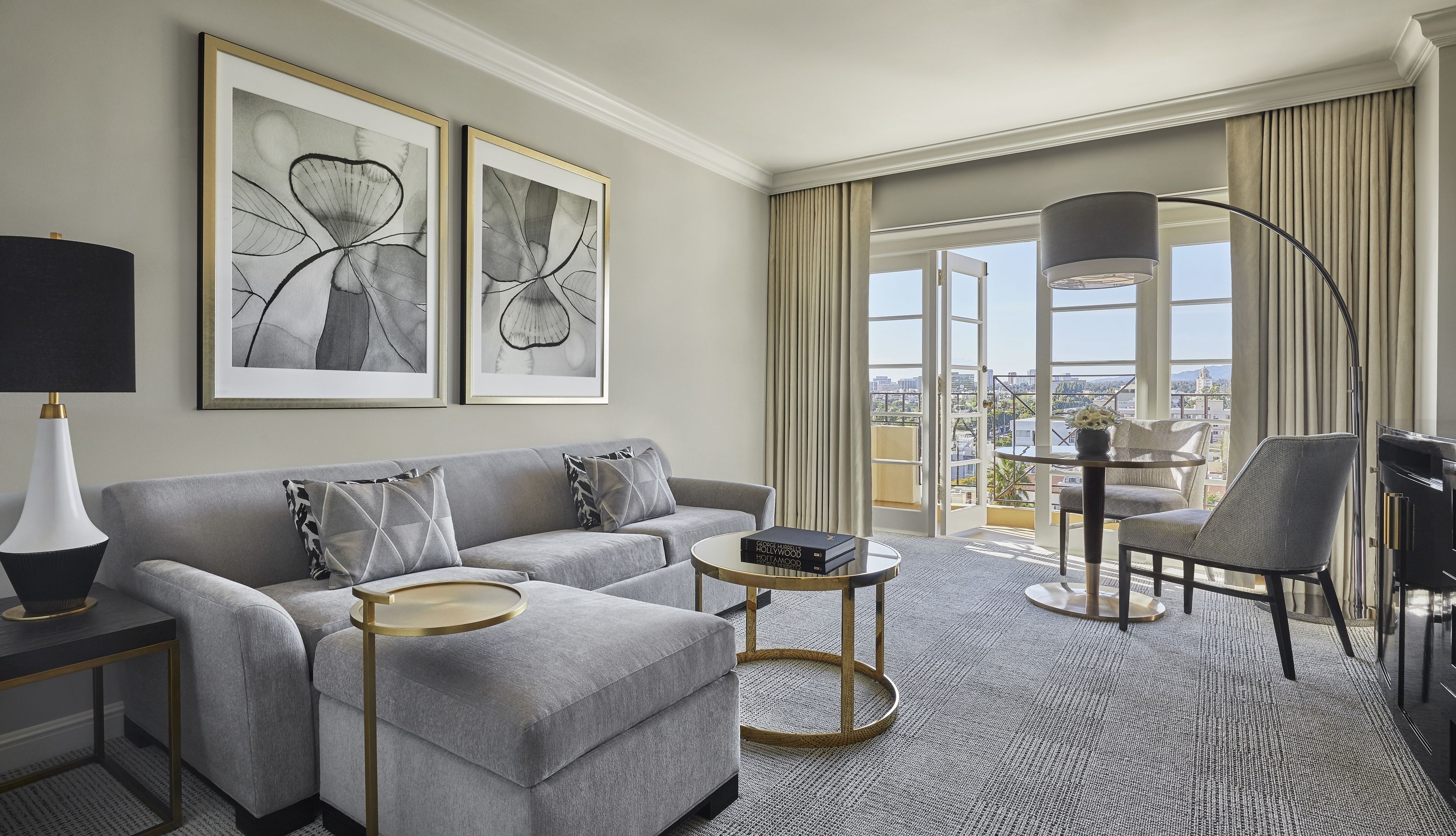 A modern living room with a grey sofa, a round coffee table, and a bookshelf. There are two framed artworks on the wall, a floor lamp, and a view of the city through large windows.