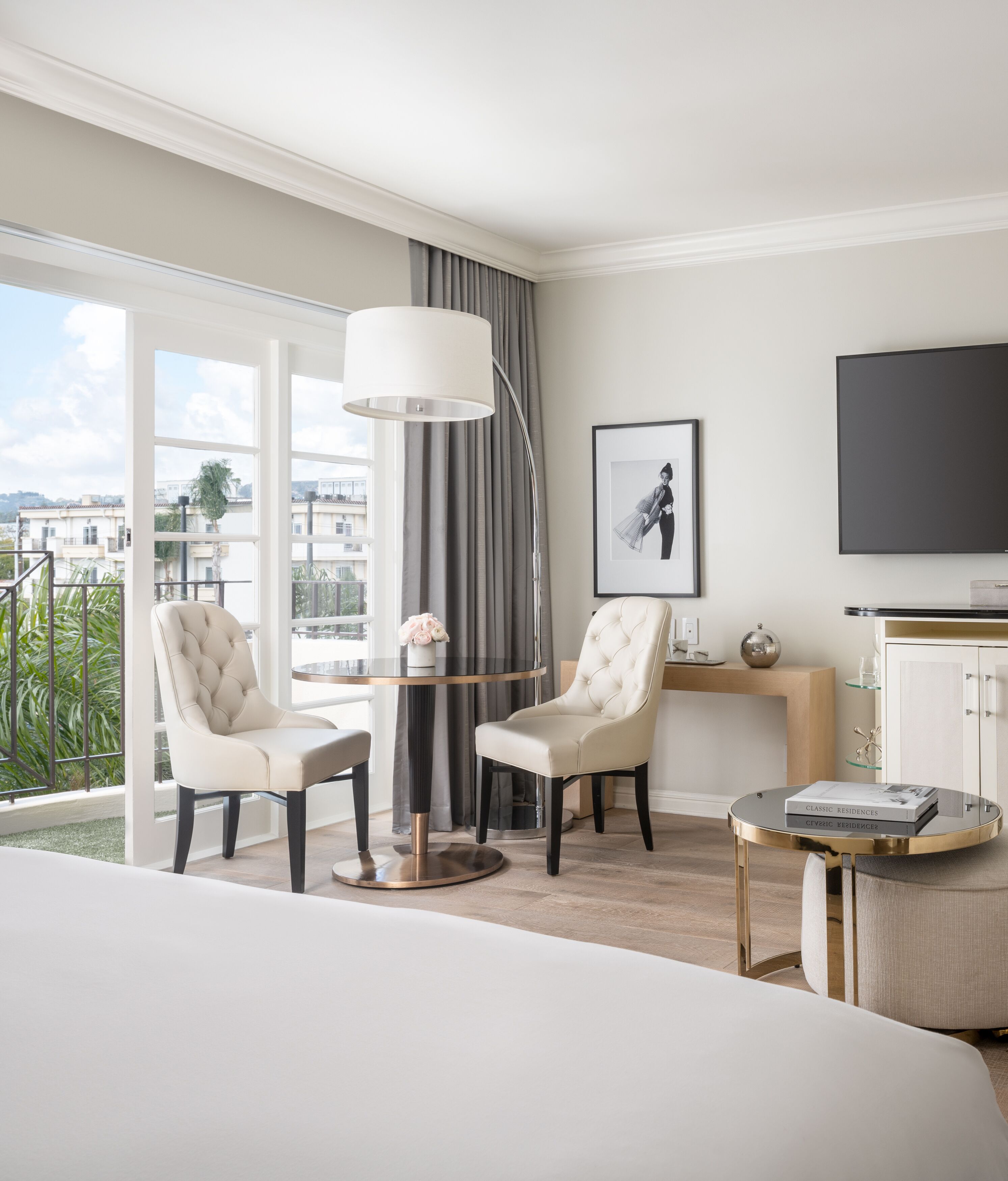 Ein modernes Hotelzimmer mit einem großen Bett, zwei Sesseln, einem runden Tisch, einem Fernseher und einem Balkon mit Blick auf Gebäude und Grünflächen.