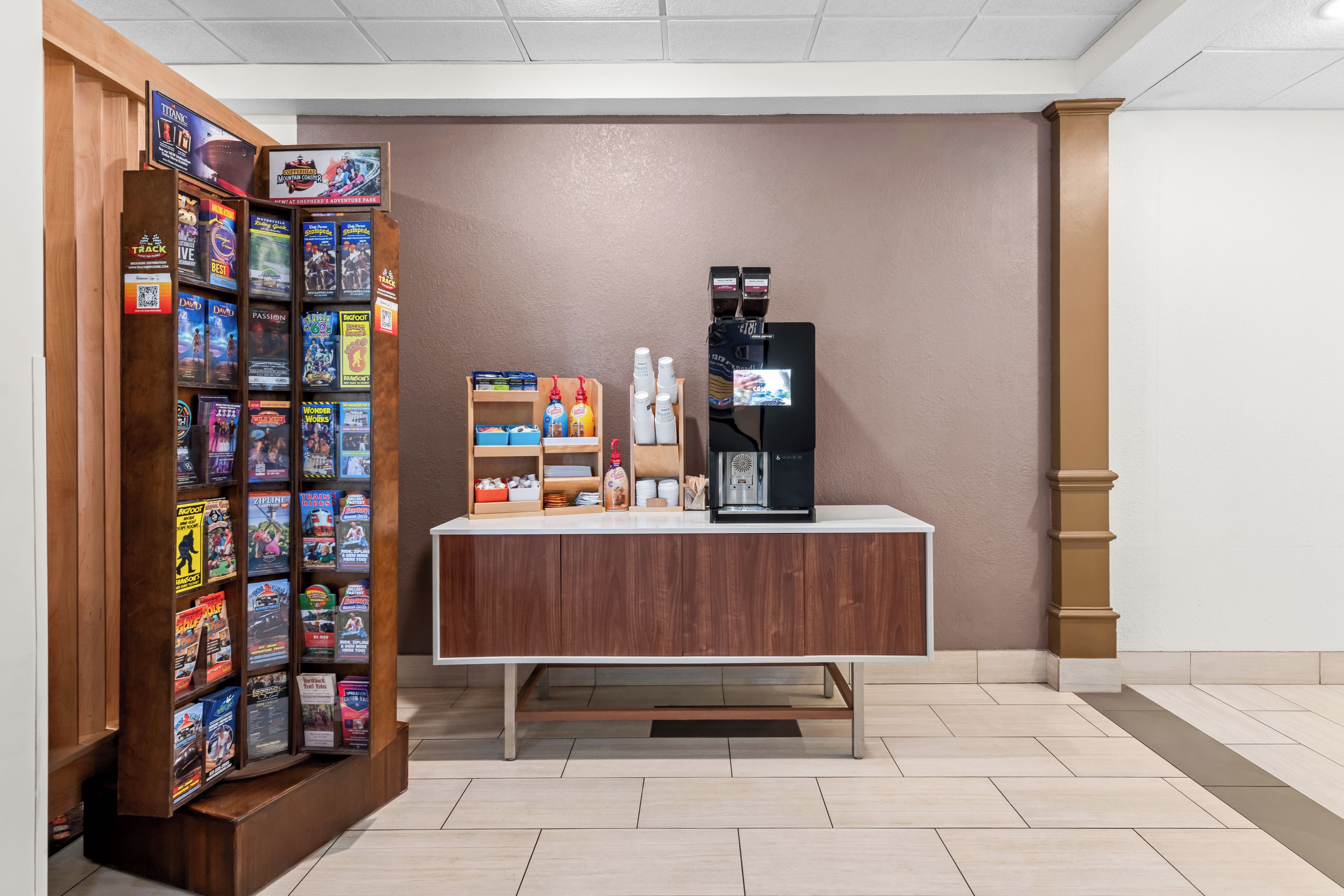 Un coin café équipé d’une machine à café et de tasses, un présentoir à magazines contenant diverses publications, et une petite étagère avec des tasses à café et une bouteille.