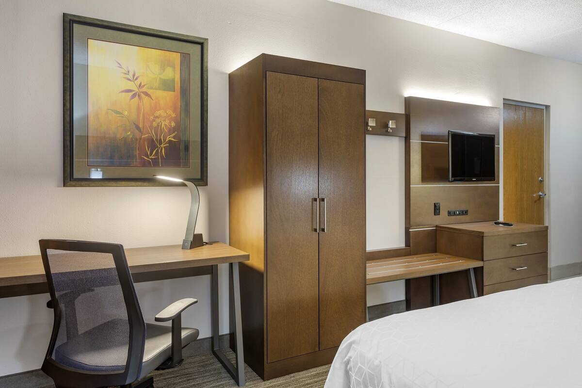 Quarto de hotel com cama, escrivaninha com cadeira, guarda-roupa, televisão e quadro na parede.