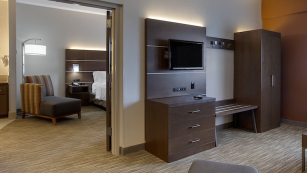 A hotel room with a flat-screen TV mounted on the wall, a desk with drawers, a large wardrobe, a cushioned chair, and a bedside table with a lamp. - Holiday Inn Express Branson-Green Mountain Drive by IHG (Branson)