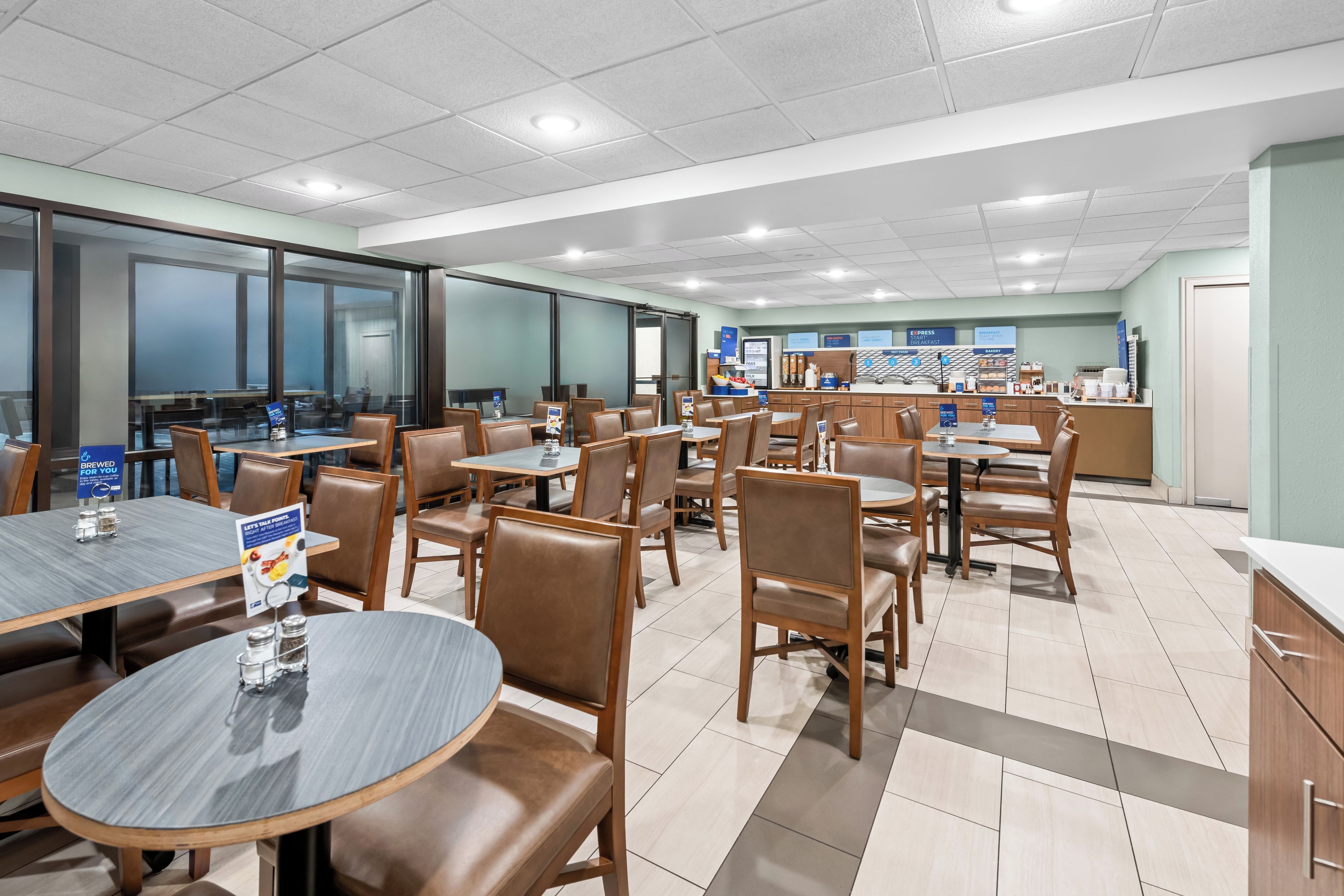 A modern dining area with multiple tables and chairs, a kitchen area with a coffee station, and a view through the glass wall.