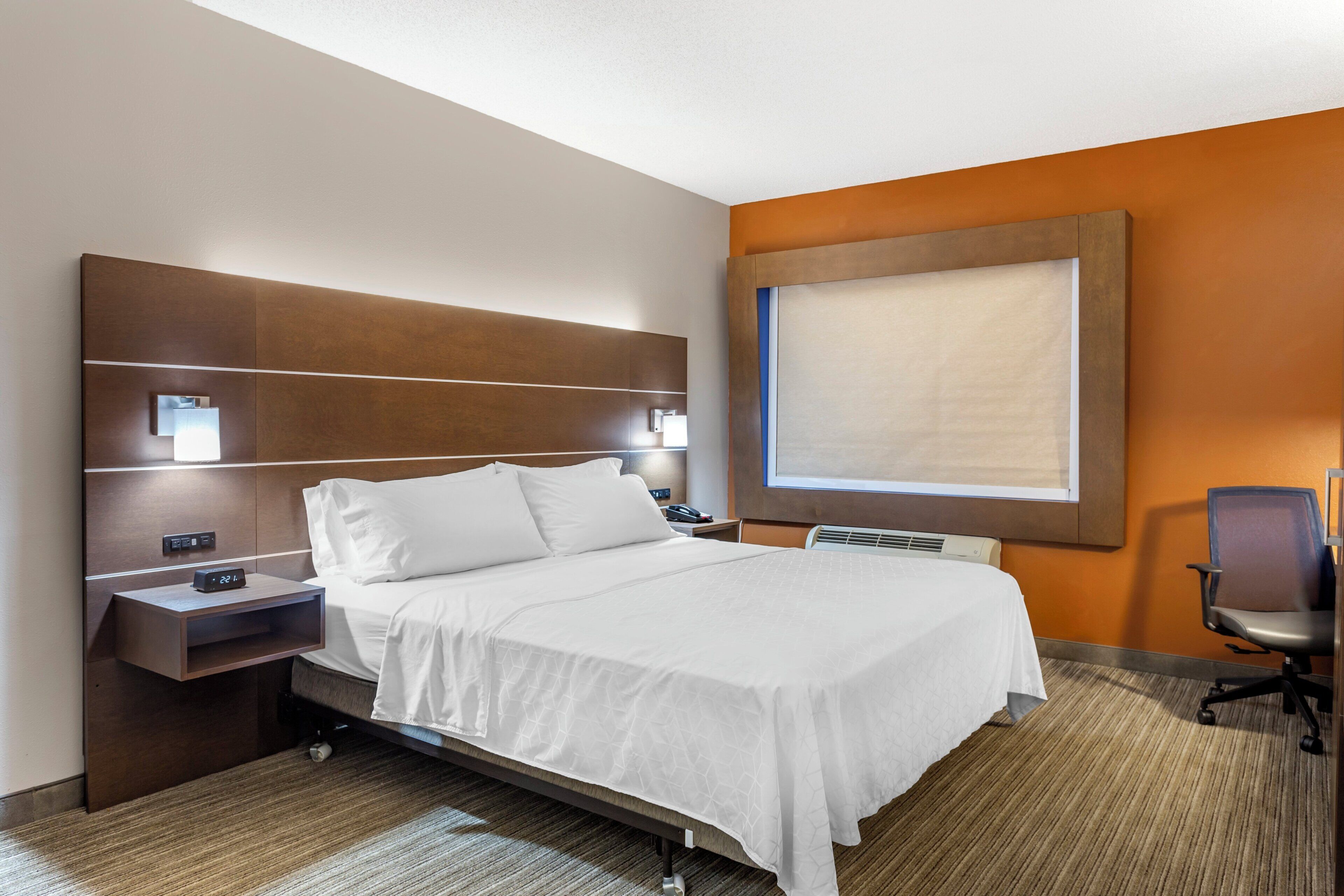 A hotel room with a bed, bedside tables, a desk, a chair, and a large window.