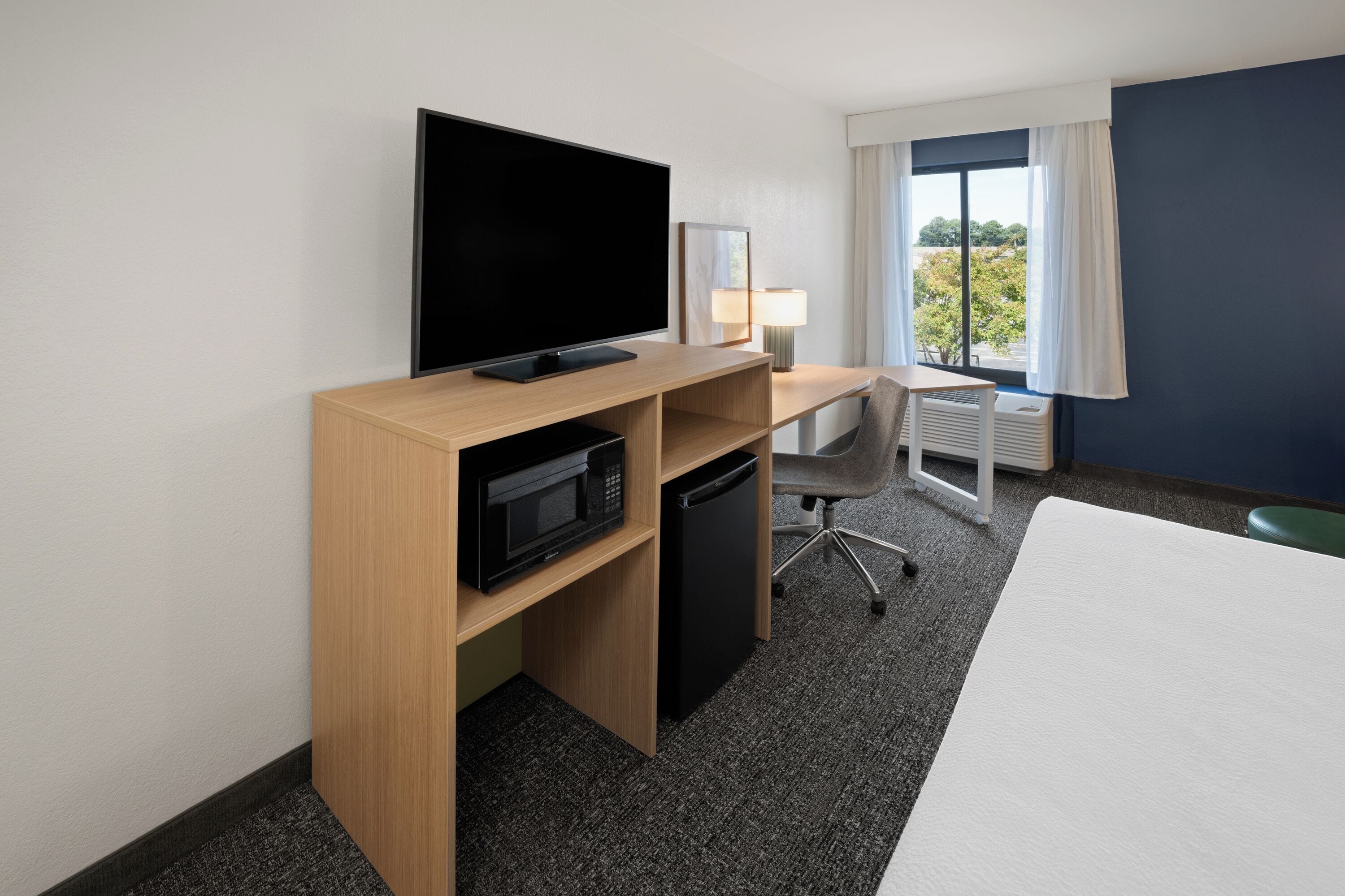 A hotel room with a flat-screen TV, a microwave, a mini-fridge, a desk with a chair, and a large bed.