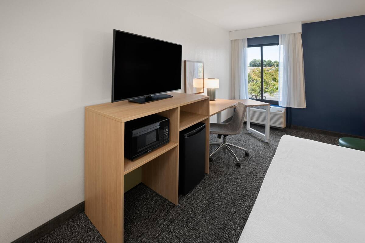 A hotel room with a bed, a desk with a flat-screen TV, a microwave, a mini-fridge, a chair, and a window with curtains.