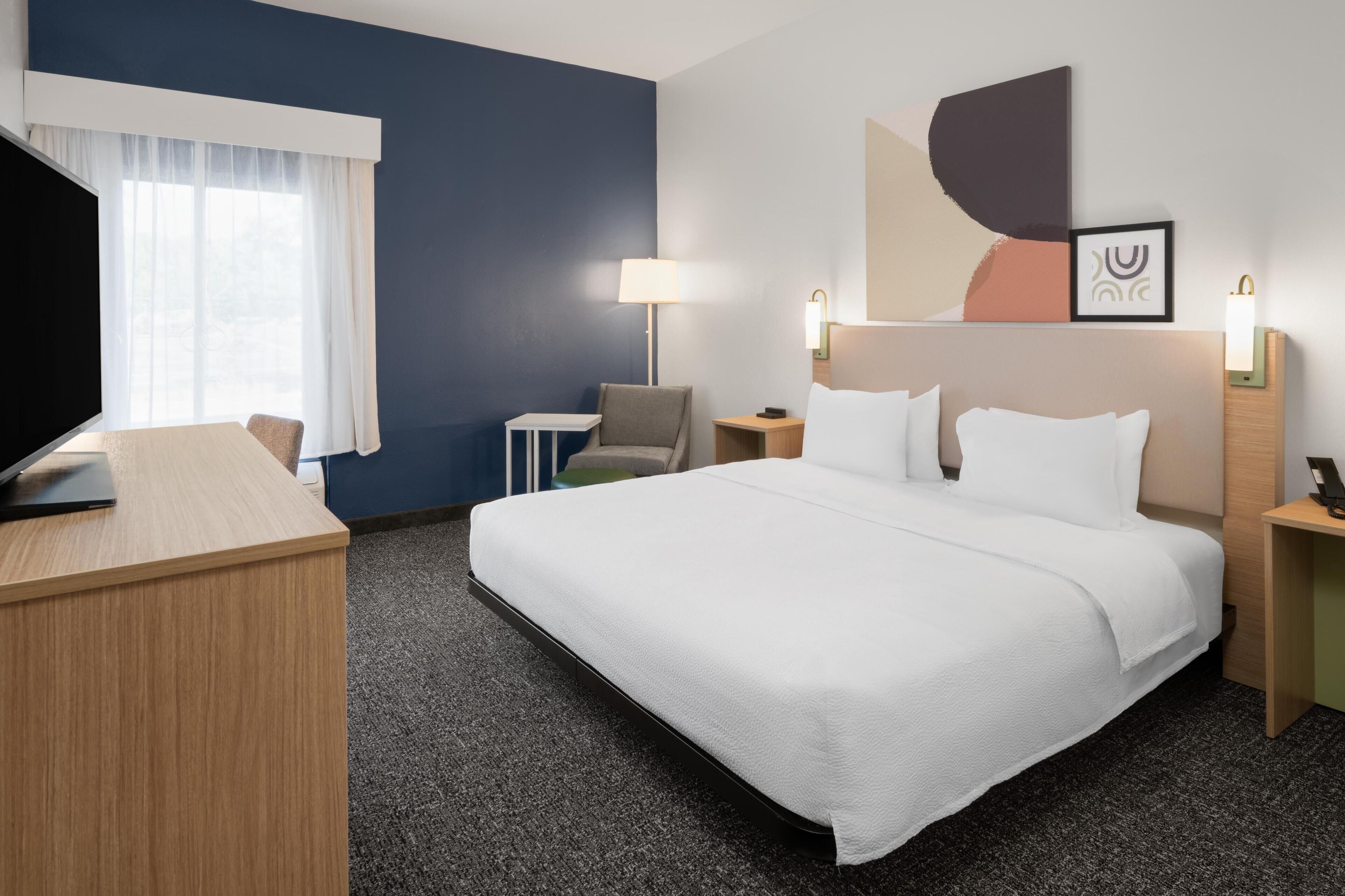 A hotel room with a bed, a desk, a chair, a TV, a lamp, and a wall with artwork.