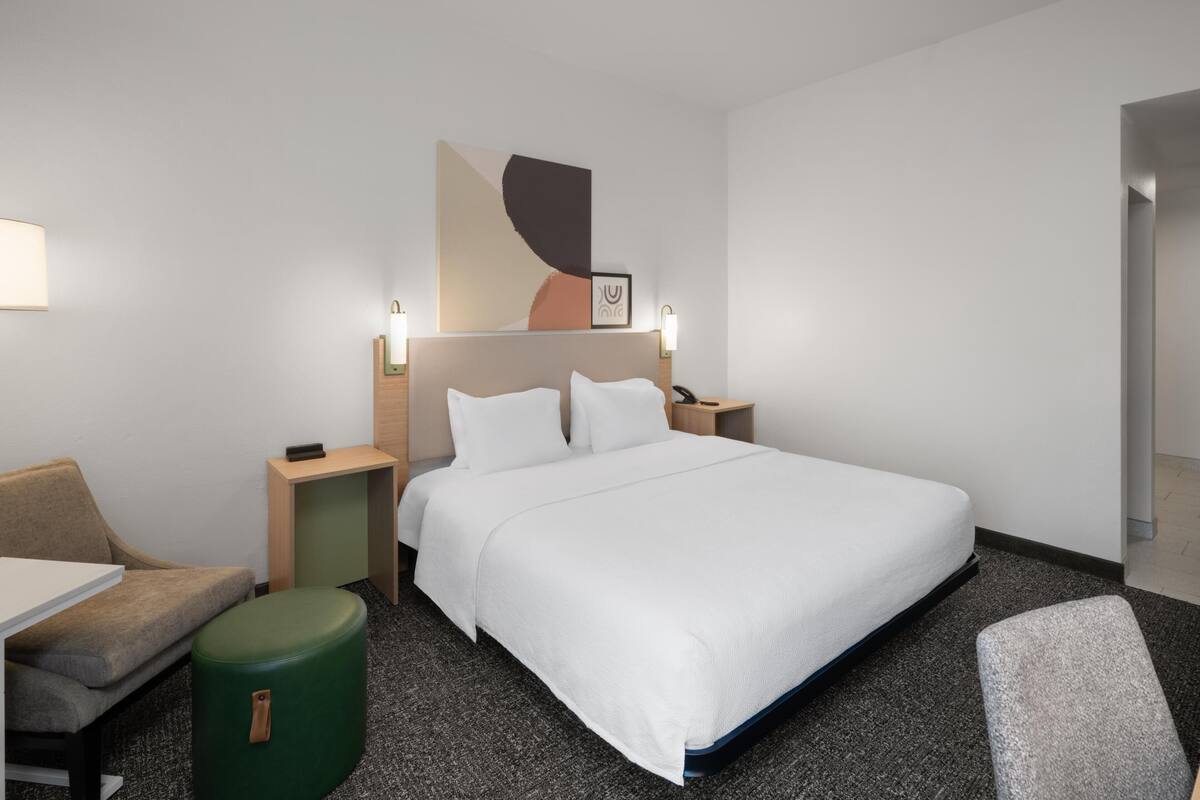 A hotel room with a bed, a desk, a chair, a green ottoman, and a wall-mounted abstract art piece.