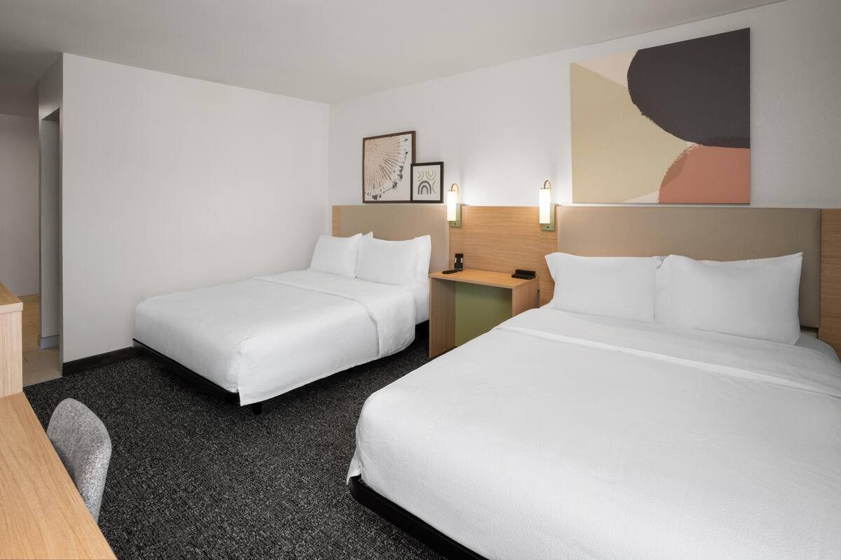 A hotel room with two beds, a desk, a chair, and a nightstand with a lamp.
