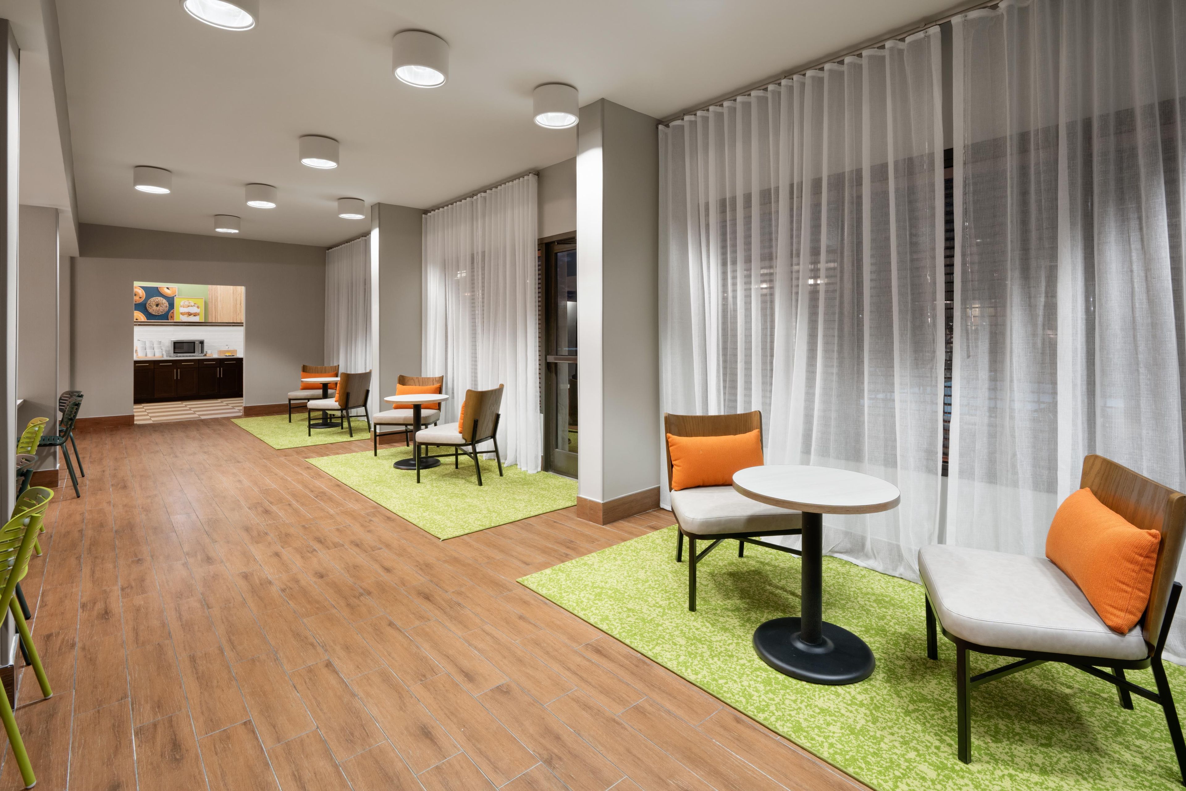 A modern hotel lobby with a dining area, a kitchen, and a large window with sheer curtains.