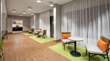 A modern hotel lobby with a dining area, a kitchen, and a large window with sheer curtains.