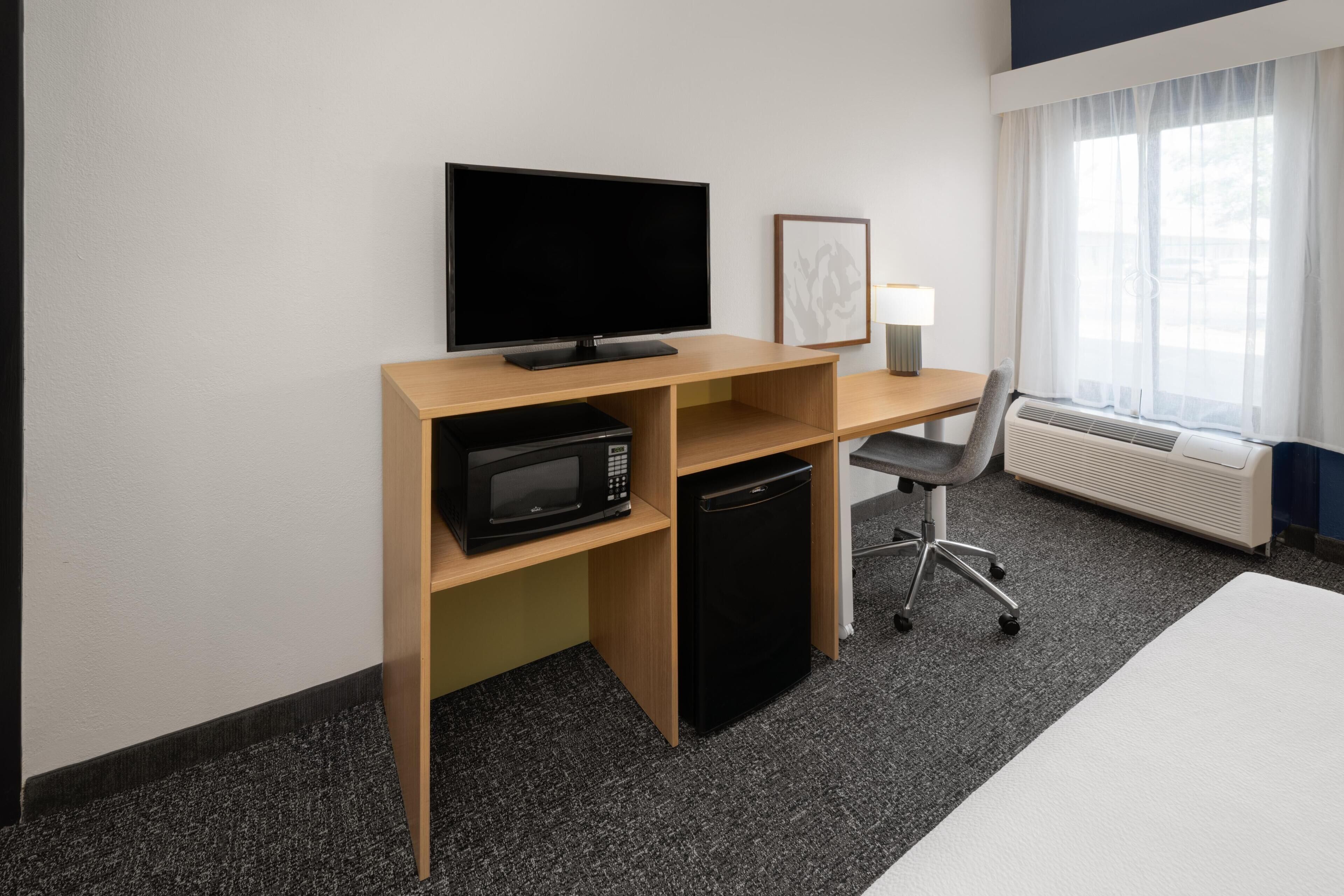 Habitación de hotel con televisión de pantalla plana, microondas, mini refrigerador, escritorio con lámpara, una silla y una cama.