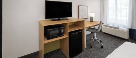 A hotel room with a wooden desk, a flat-screen TV, a microwave, a mini-fridge, a chair, a bedside table, and a large window with curtains.