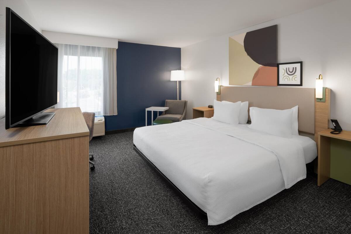 A hotel room with a large bed, a TV, a desk, a chair, a lamp, and a bedside table.