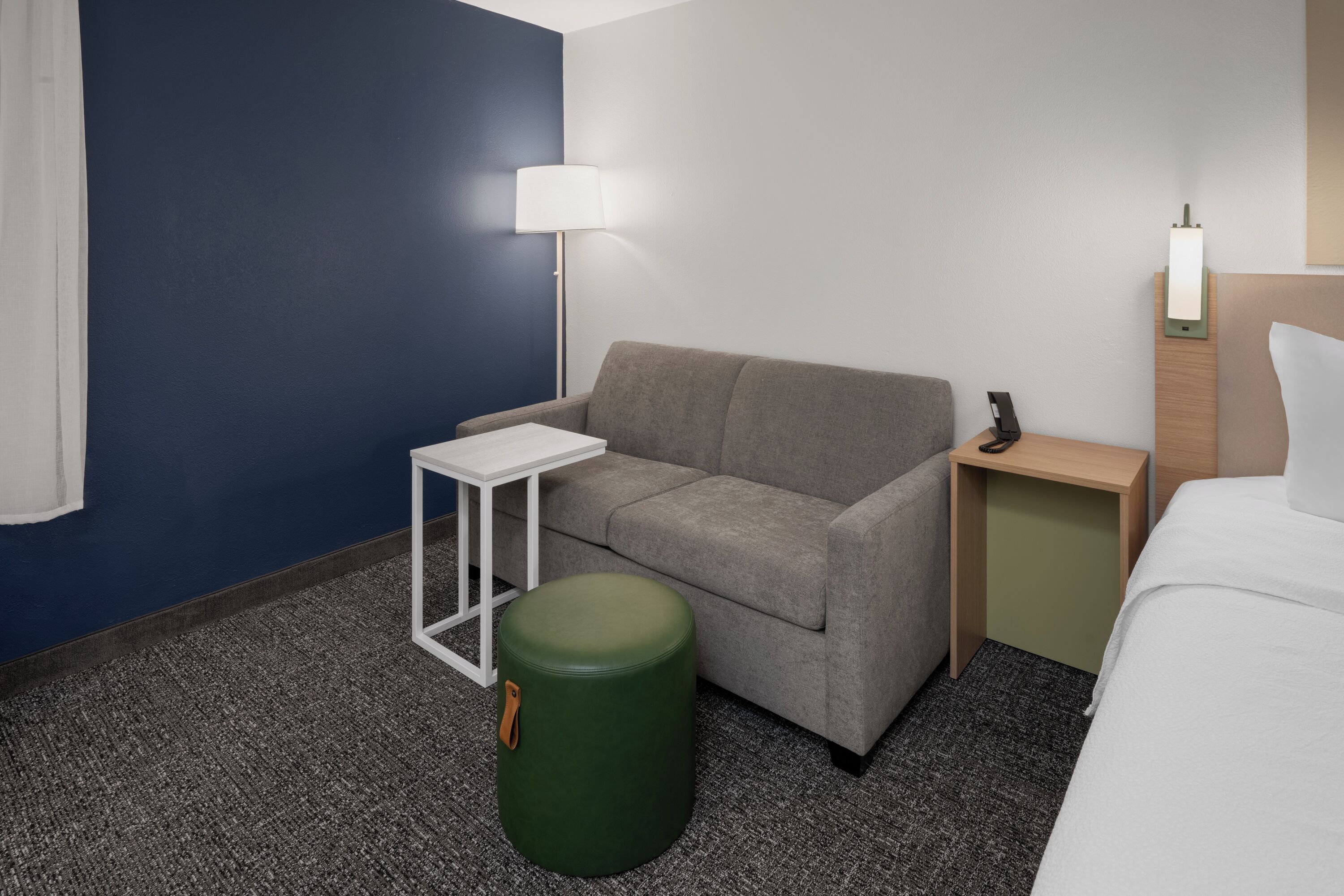 Habitació d'hotel amb un sofà gris, una taula blanca petita, un otomà verd, una taula de nit amb una làmpada i un llit amb roba de llit blanca.