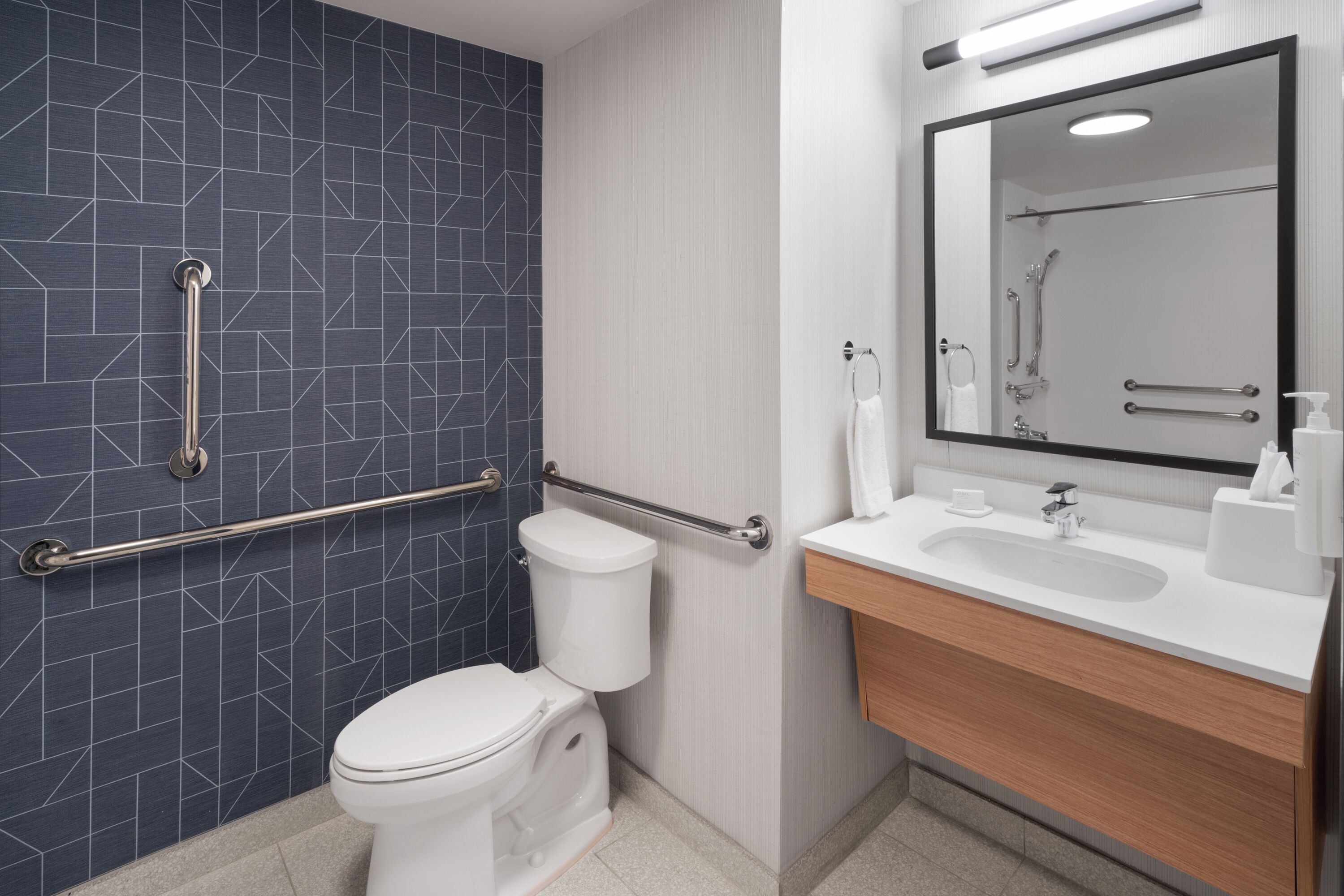 Un bany modern amb un mosaic de rajoles geomètric, un vàter blanc, un lavabo amb mirall i barres de sujeció.