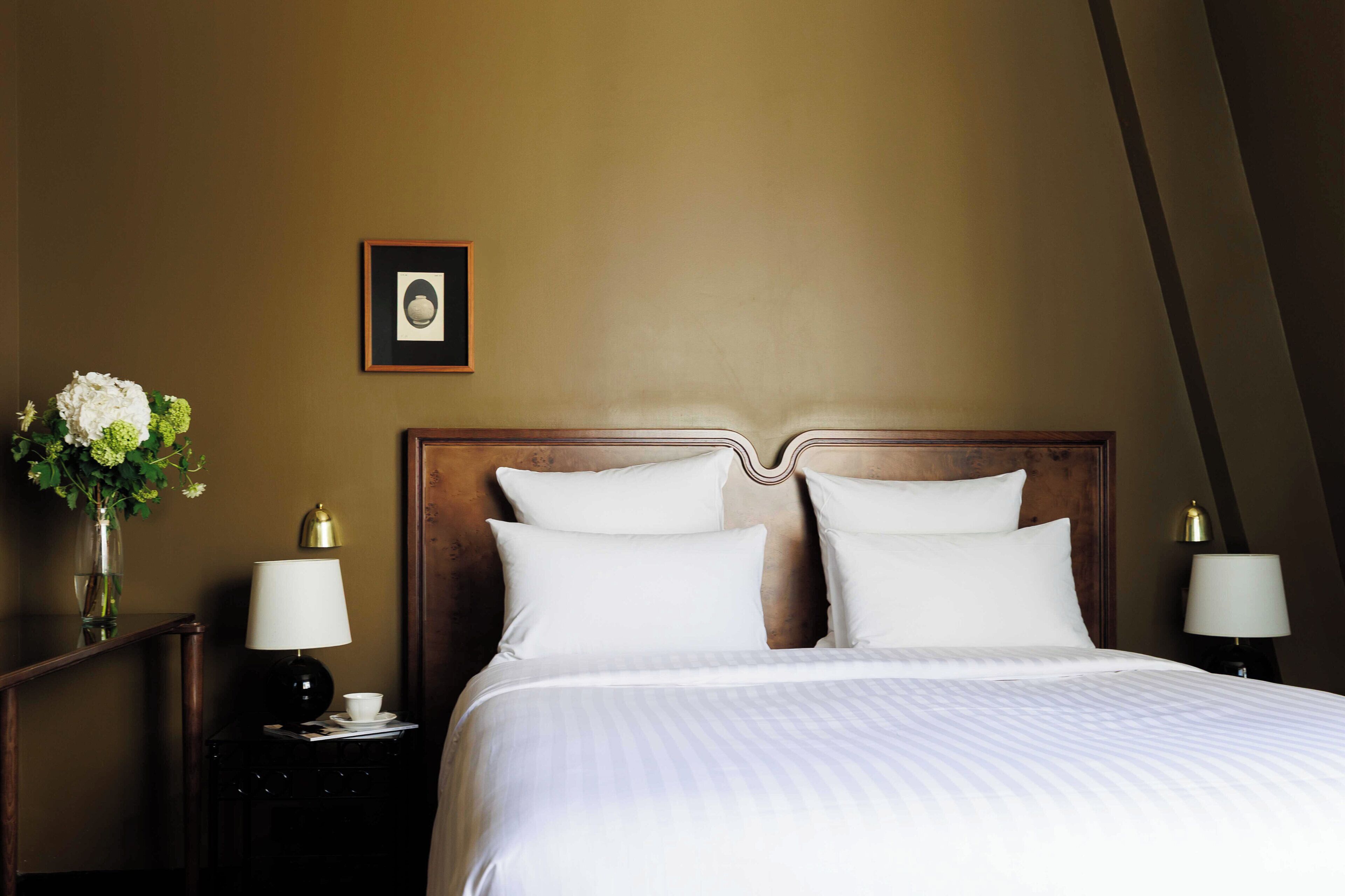 Une chambre d’hôtel avec un grand lit, deux tables de chevet, une petite table, un tableau encadré au mur et un vase avec des fleurs.