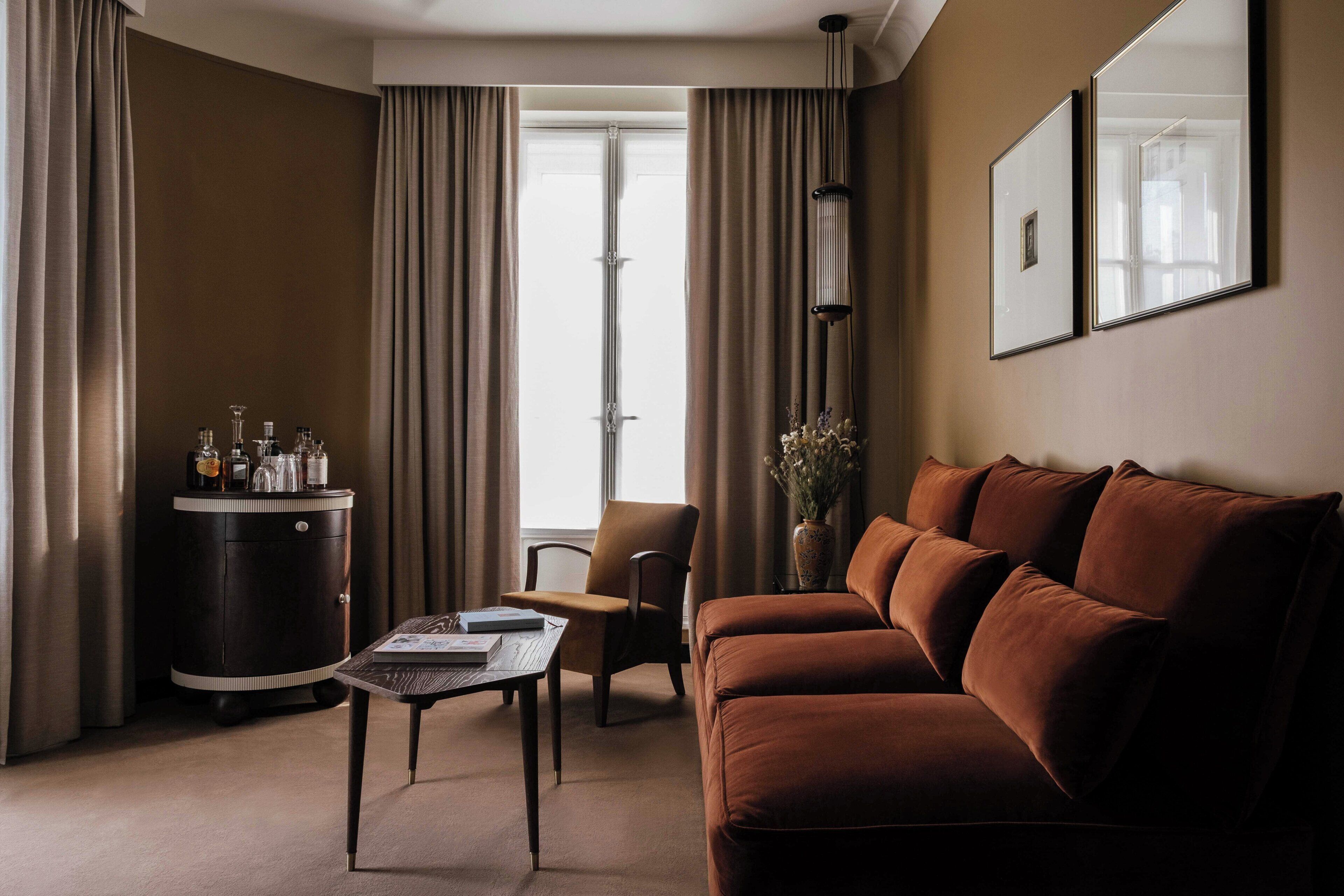 A modern living room with a brown sofa, a small table, a chair, a cabinet with bottles, and a vase with flowers.