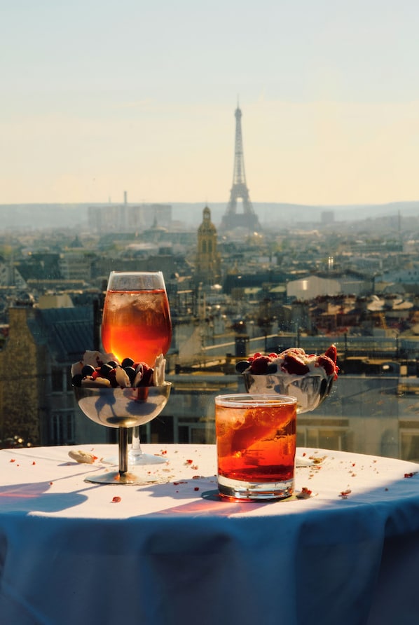 Uma mesa com dois copos de bebida vermelha e uma tigela de frutas vermelhas, com uma vista da cidade ao fundo, incluindo a Torre Eiffel.