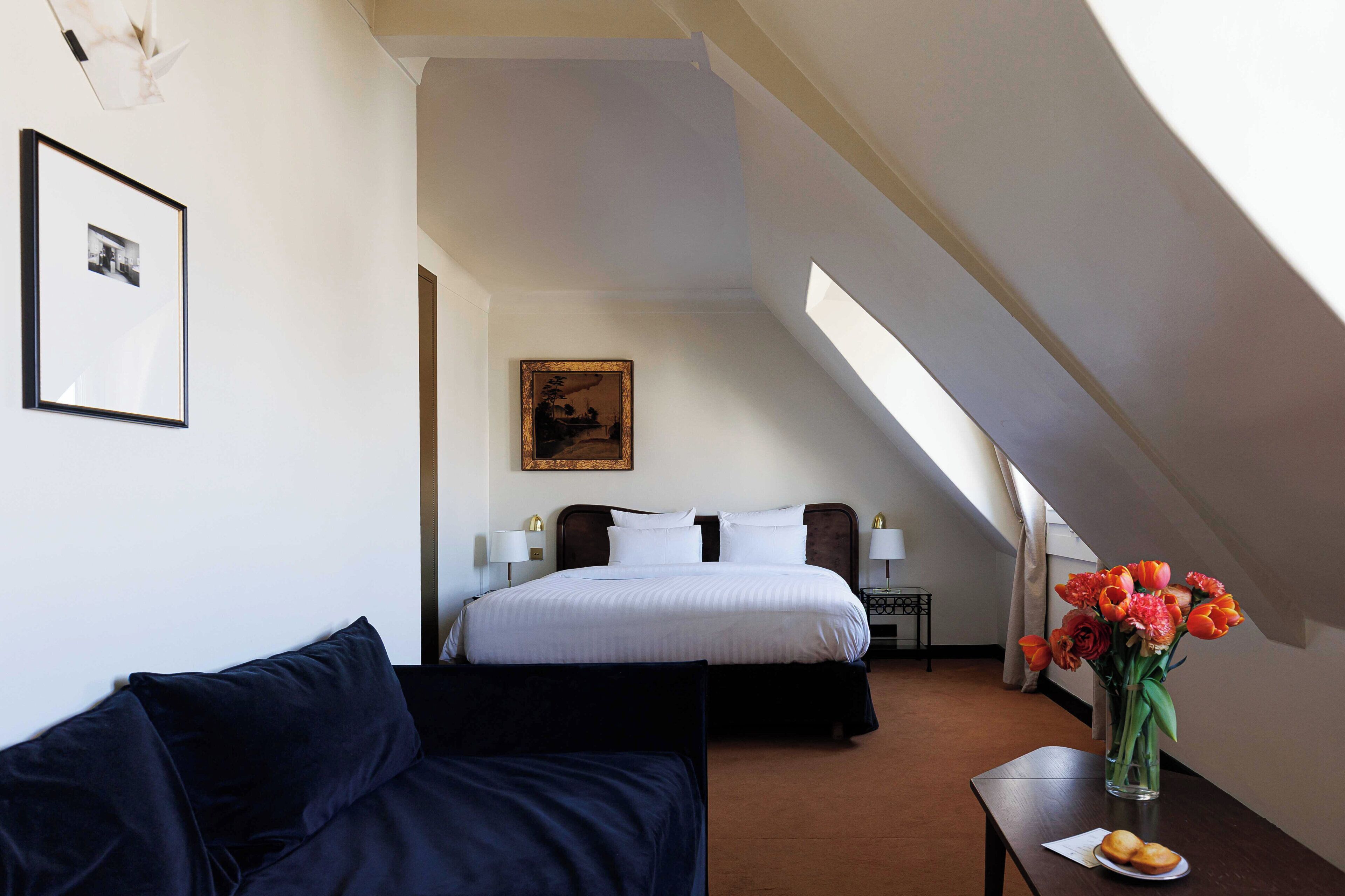 Ein Hotelzimmer mit Bett, Nachttisch, Lampe, gerahmtem Bild an der Wand, einem dunkelgrauen Sofa und einer Vase mit Blumen auf einem kleinen Tisch.