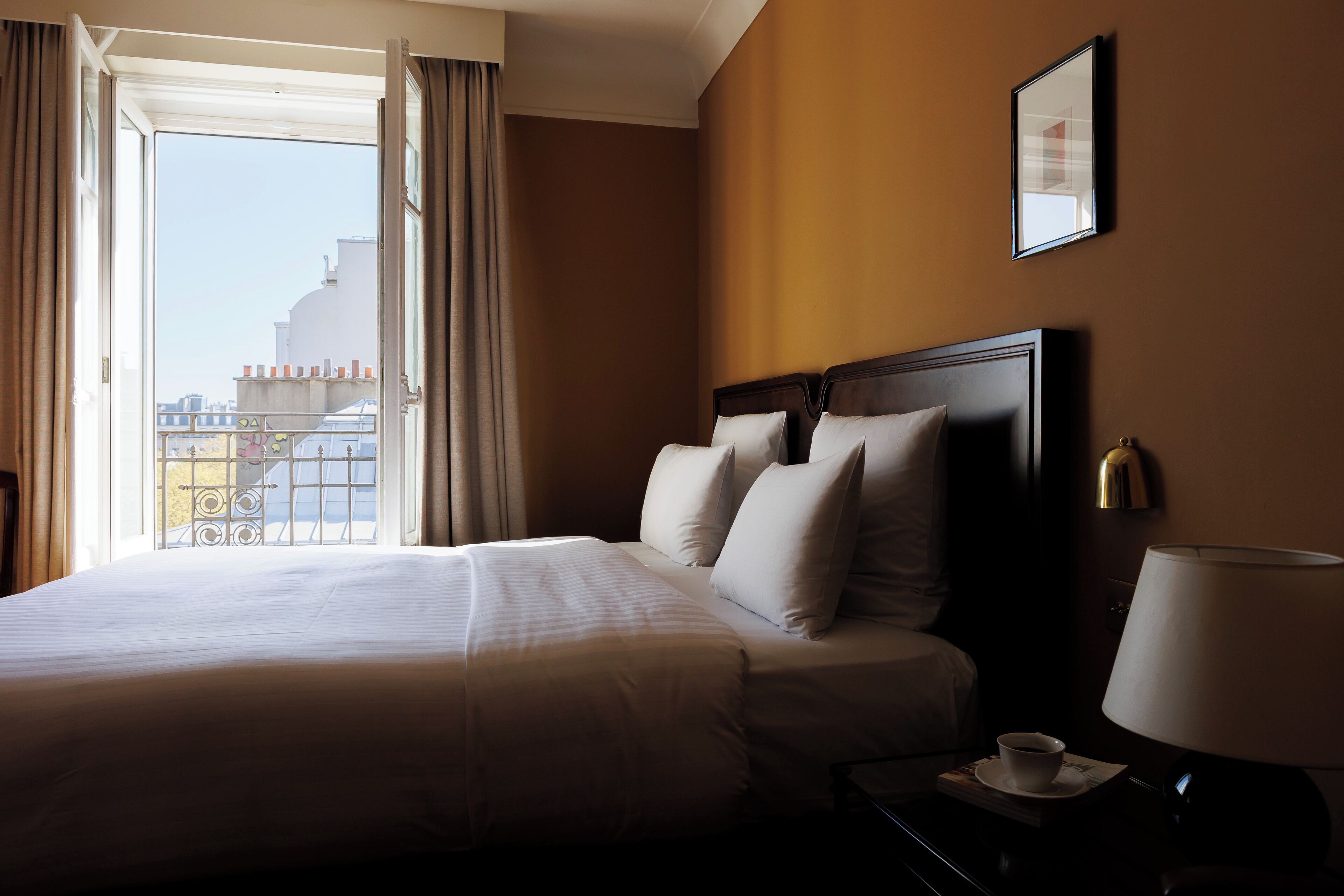 Quarto de hotel com uma cama grande, uma mesa de cabeceira com abajur e vista da cidade pela janela aberta.
