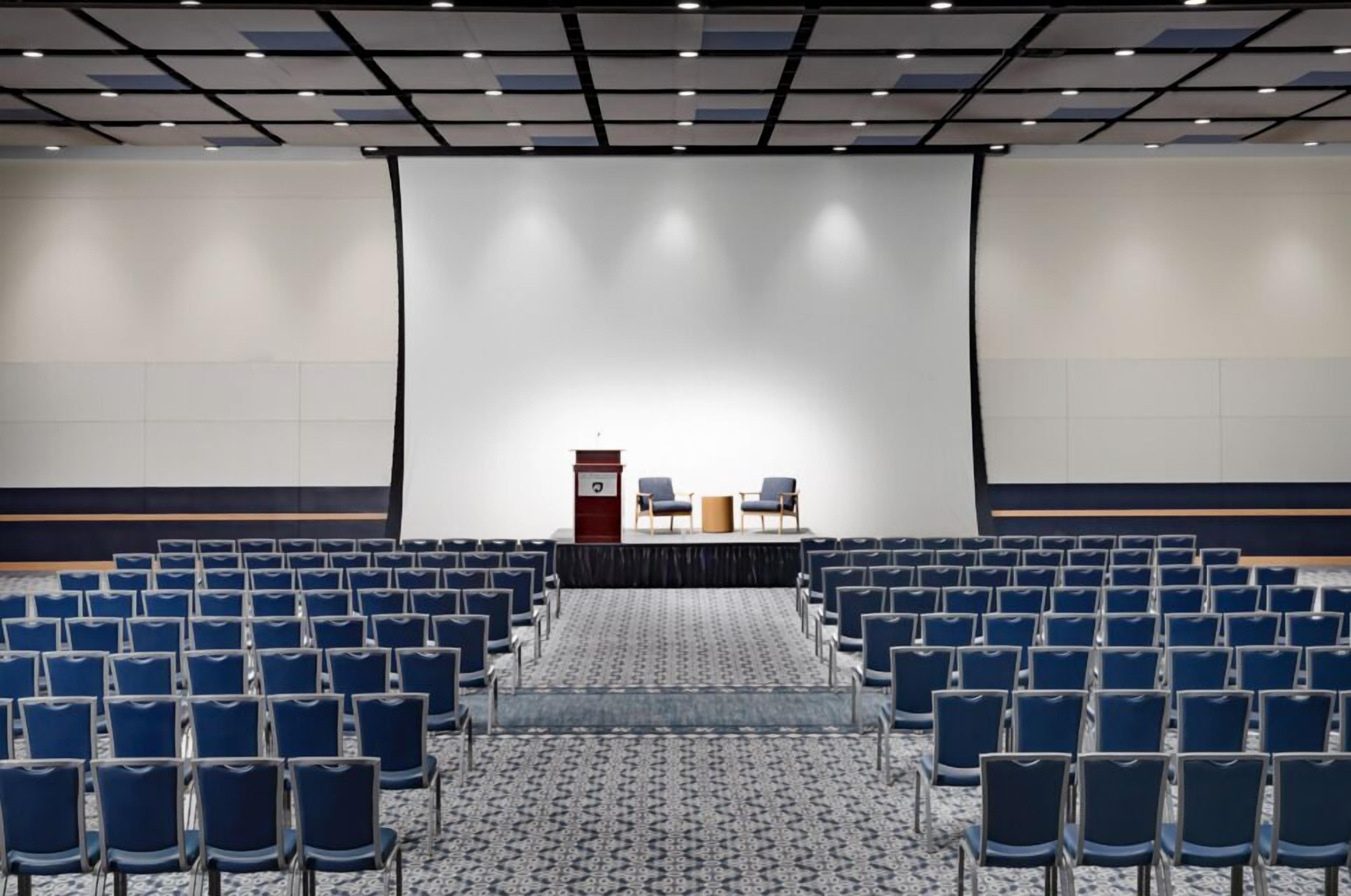 A conference room with rows of blue chairs, a podium, and a large screen at the front.