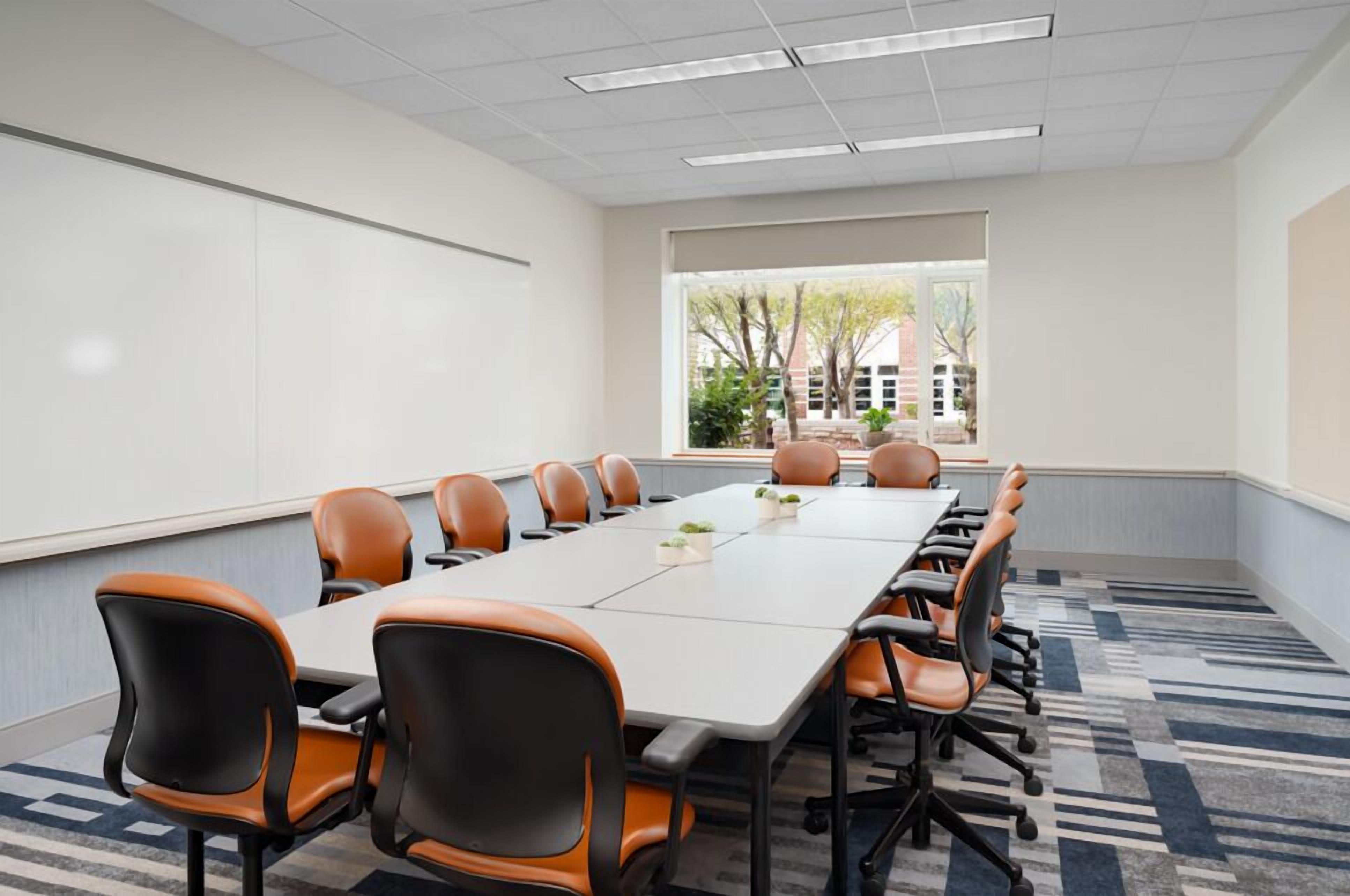 A conference room with a long table, multiple chairs, a whiteboard, and a view of trees and buildings through the window.