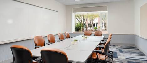 A conference room with a long table, multiple chairs, a whiteboard, and a view of trees and buildings through the window.