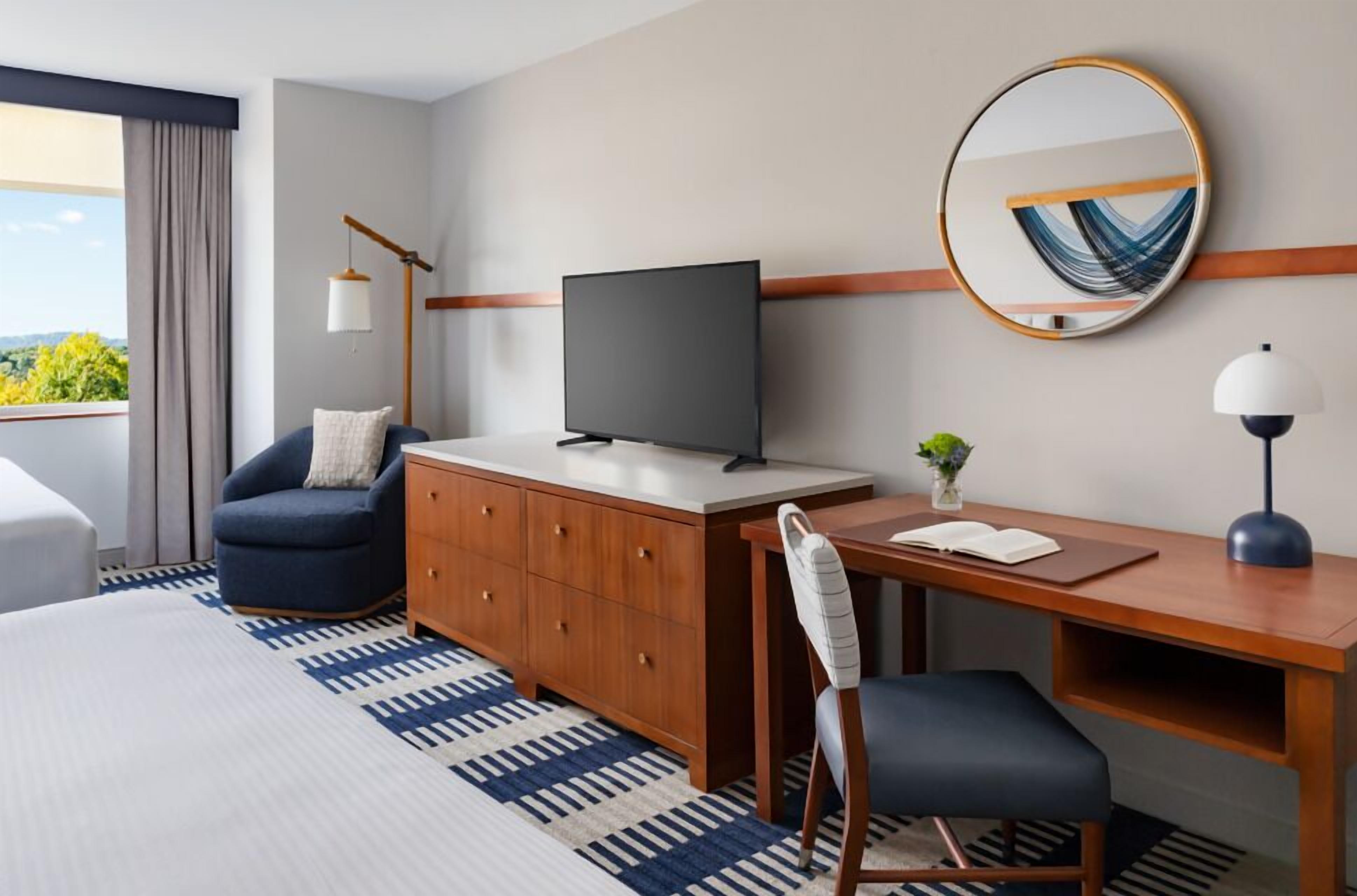 A hotel room with a bed, a desk, a chair, a television, a mirror, and a window with curtains.