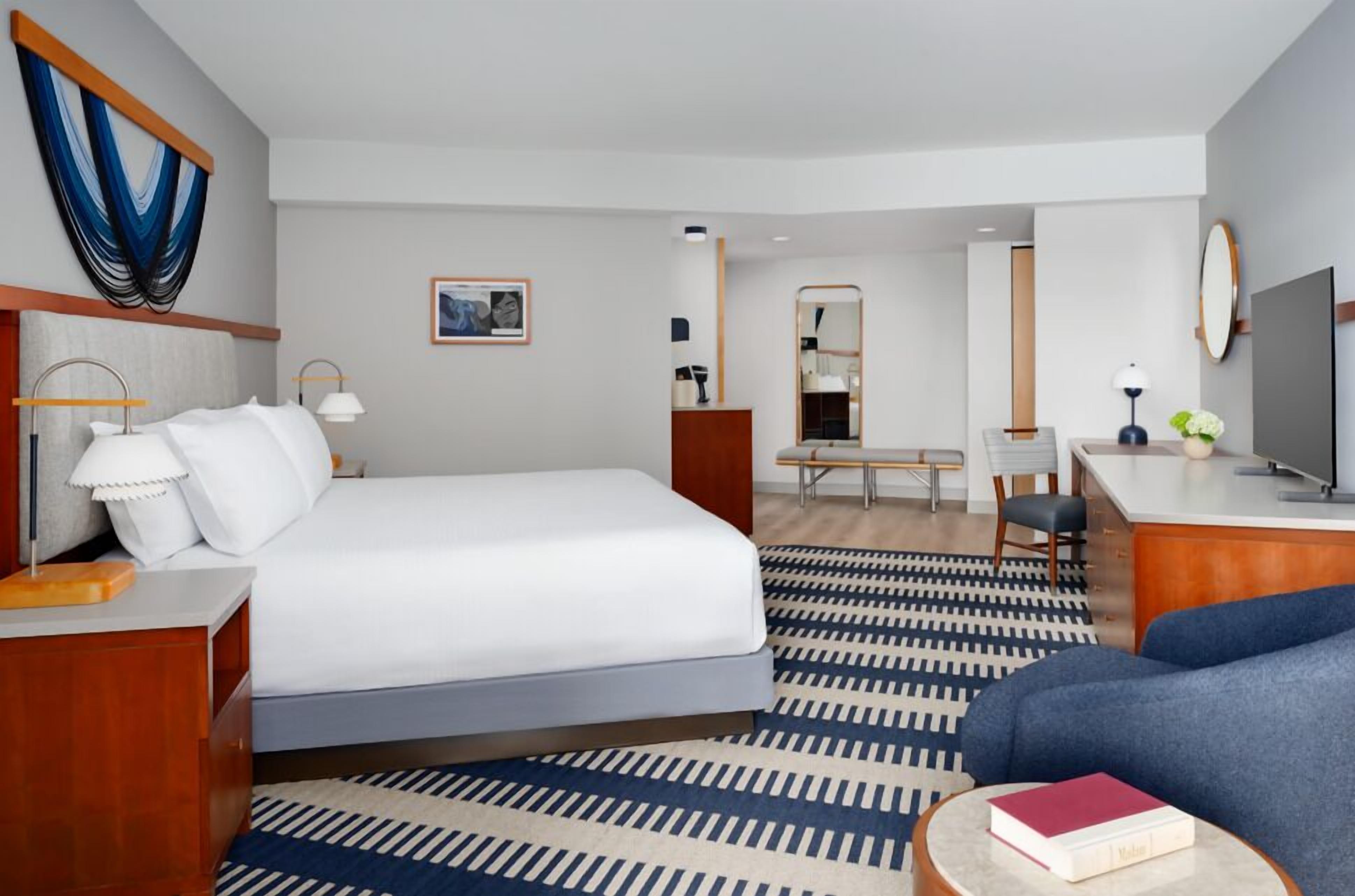Quarto de hotel com cama, mesa de cabeceira, luminária e um sofá azul.