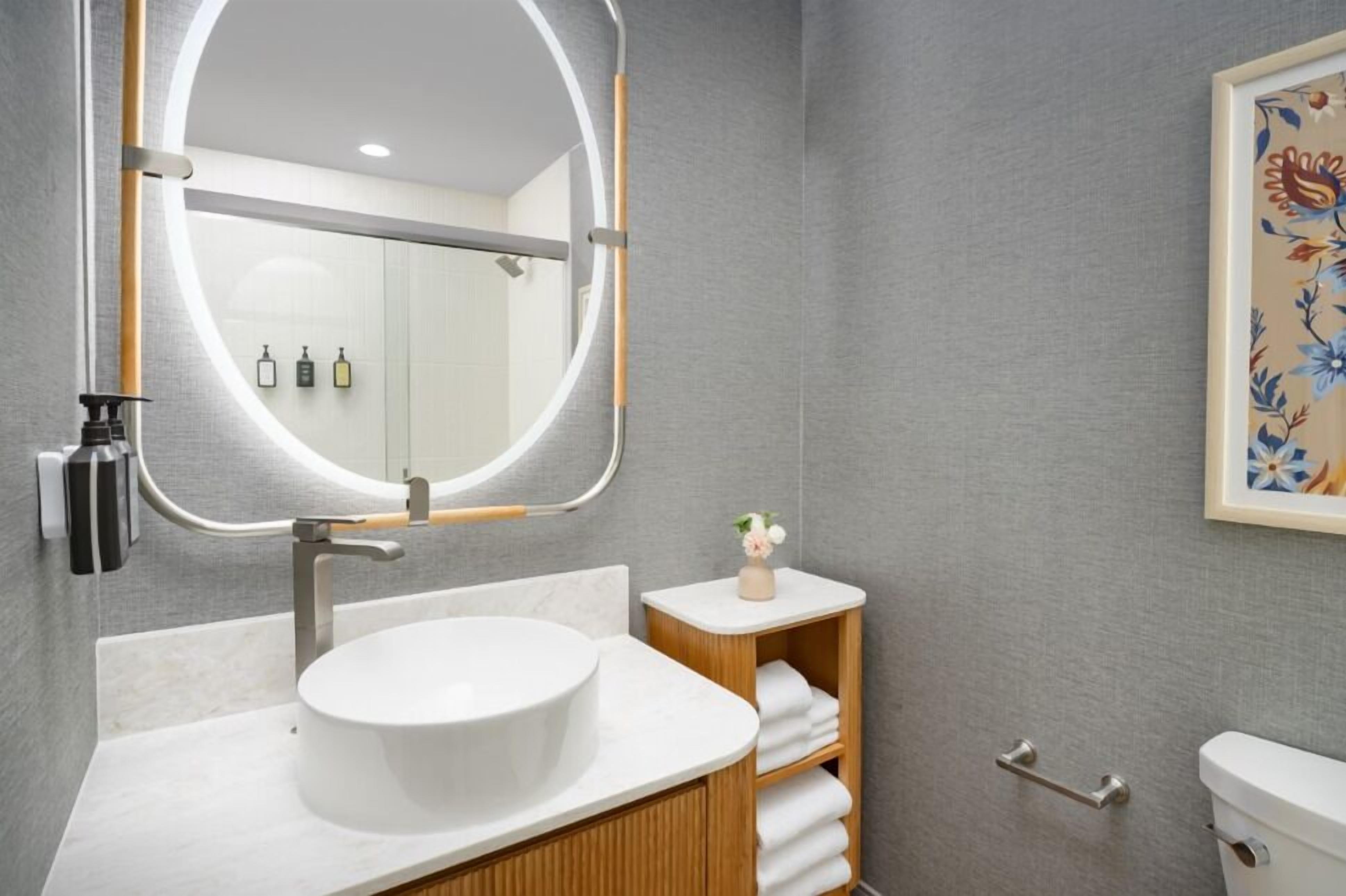 Un baño moderno con un espejo redondo grande, un lavabo blanco y un estante de madera con toallas.