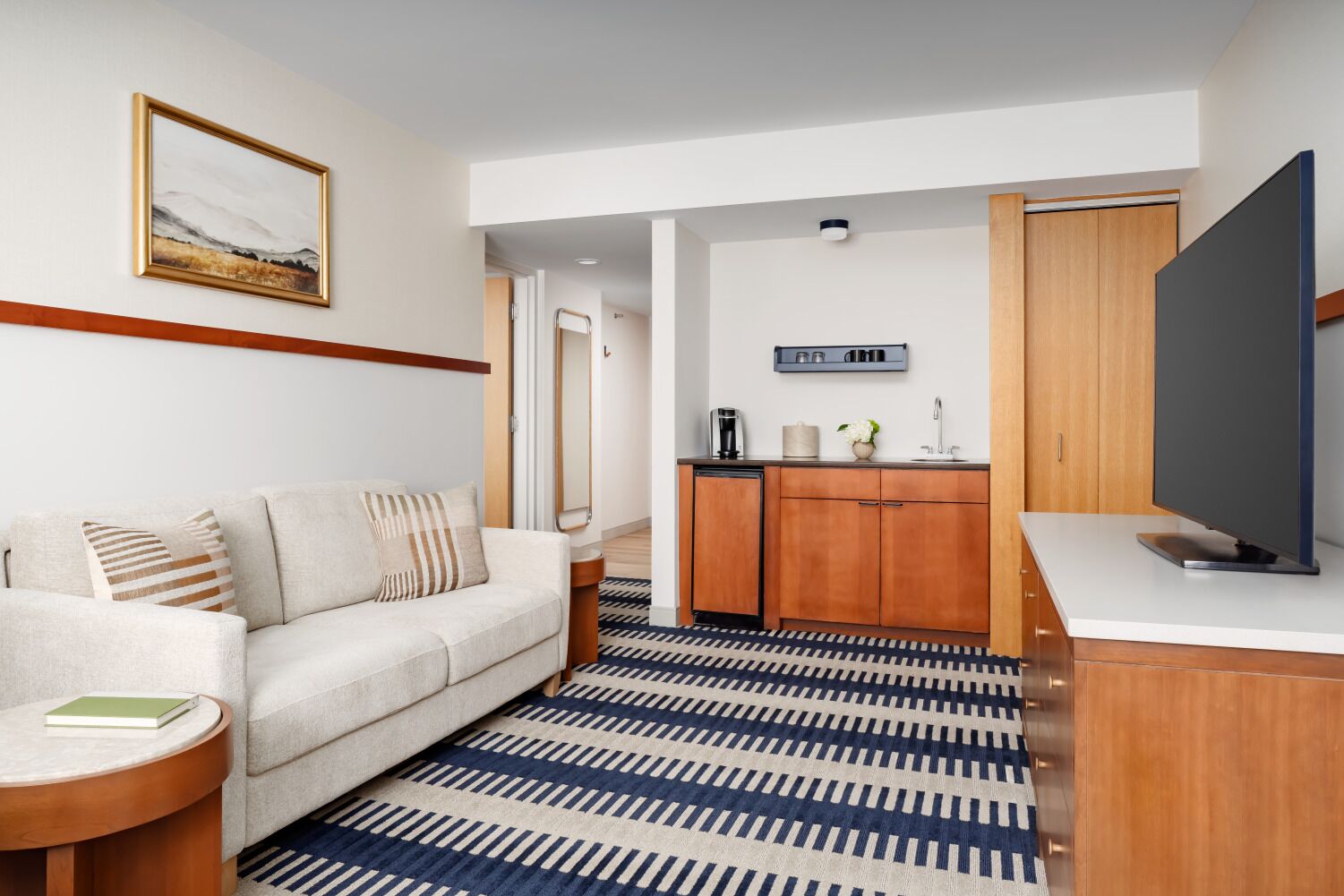 A modern hotel room with a sofa, a small side table, a coffee machine, a flat-screen TV, and a kitchen area with wooden cabinets.