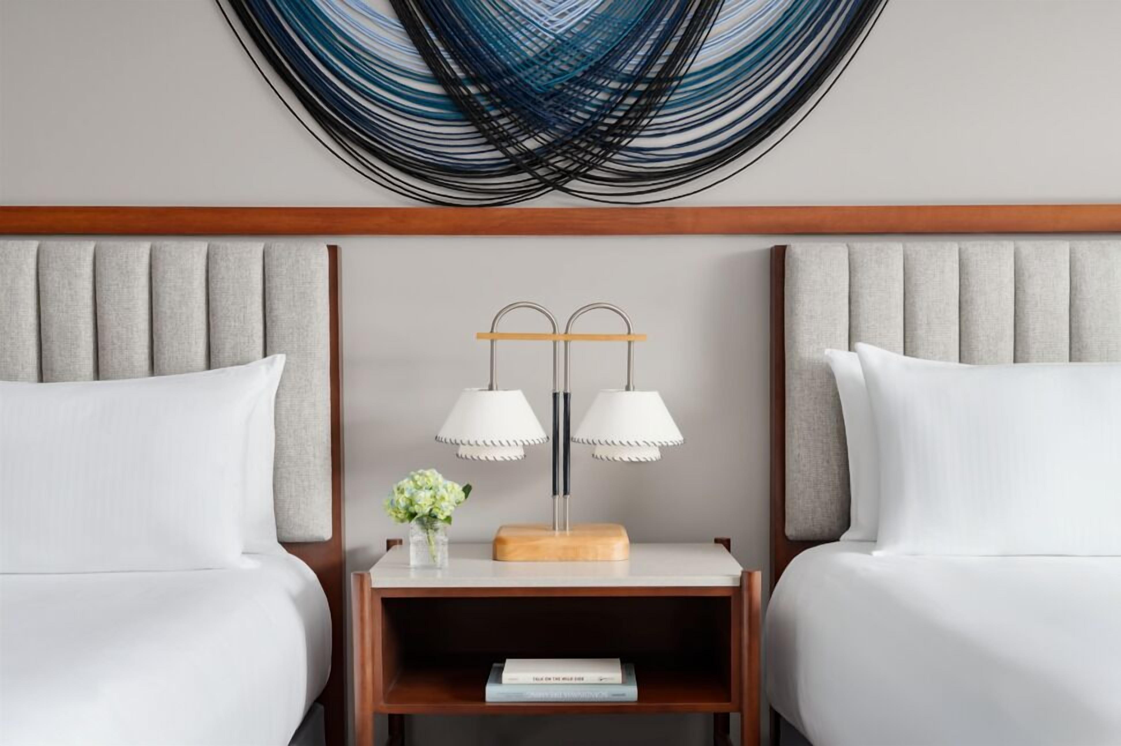 A hotel room with two beds, a wooden headboard, a nightstand with a lamp, and a wall art piece.