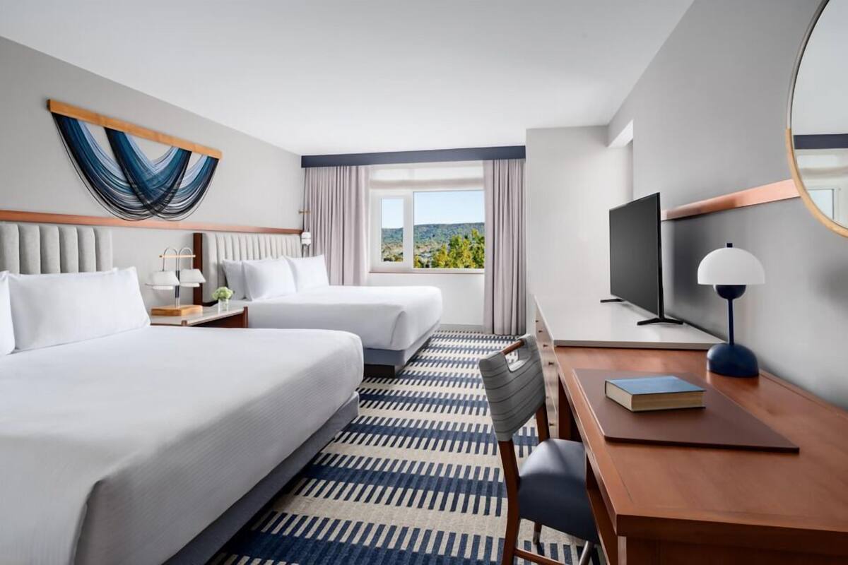 A hotel room with two beds, a desk, a chair, a TV, and a window with a view of the outdoors.