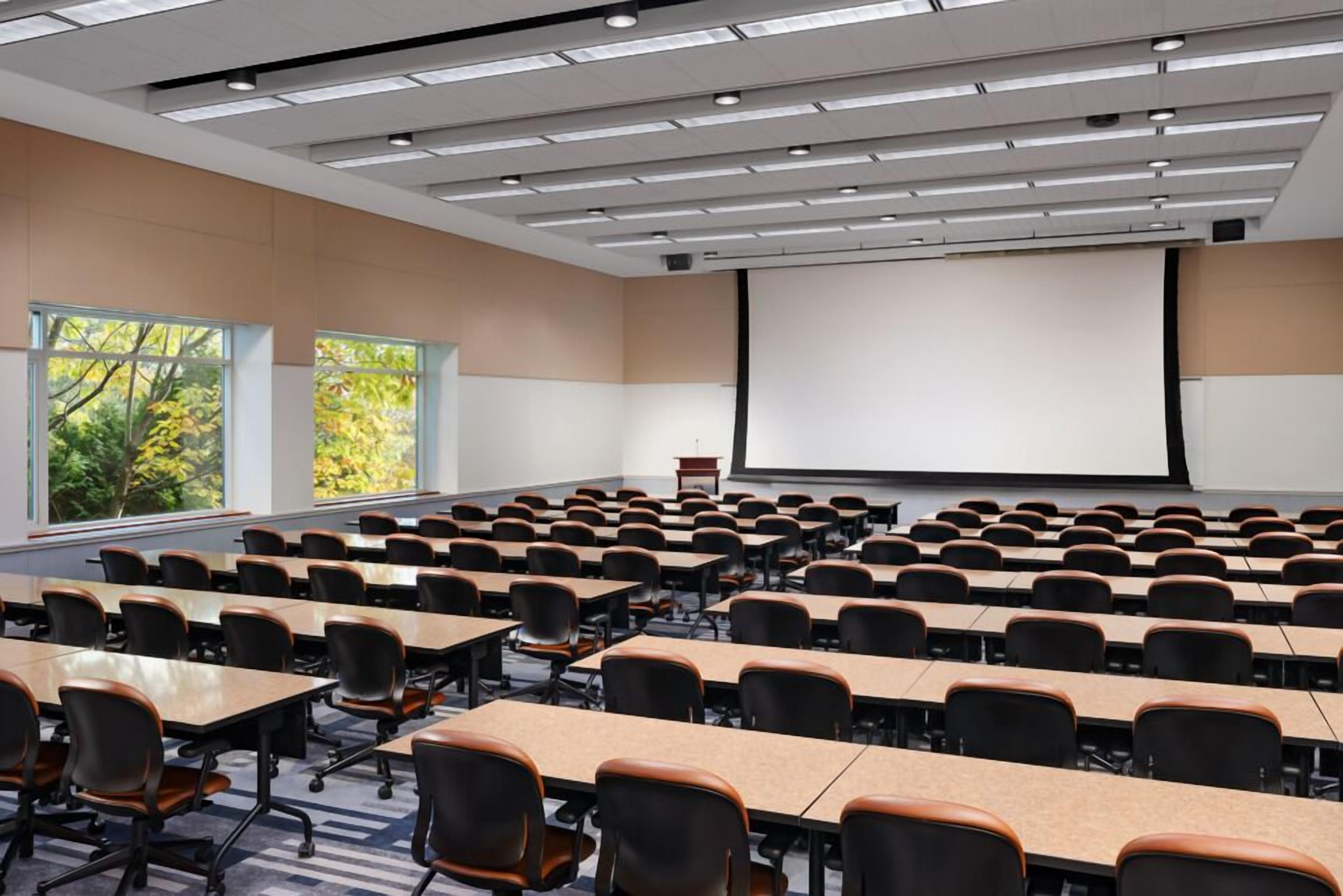 A conference room with rows of tables and chairs, large windows, and a projection screen.
