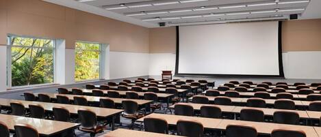 A conference room with rows of tables and chairs, large windows, and a projection screen.