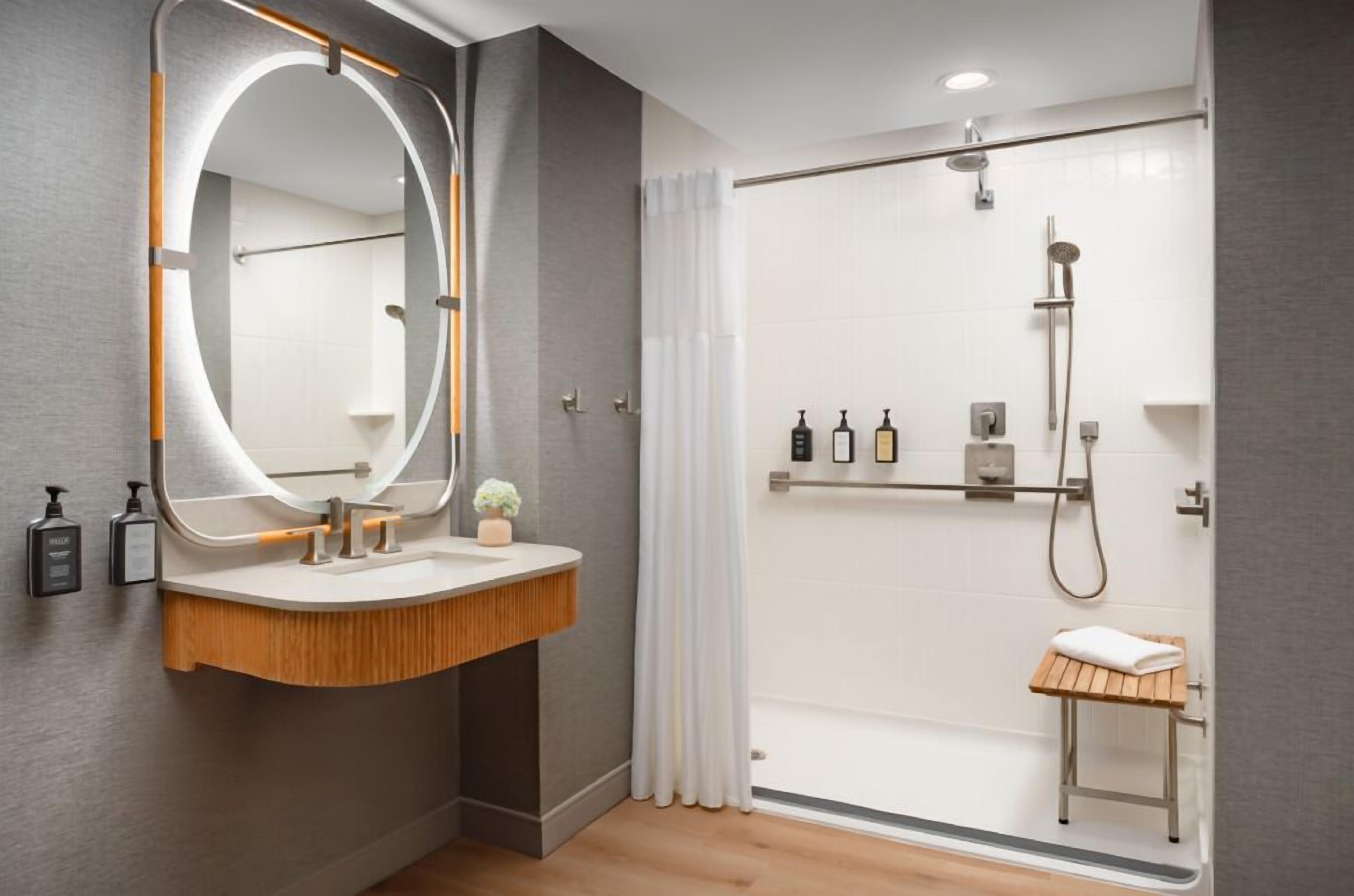 A modern bathroom with a vanity, a large mirror, a shower area, and a small stool.