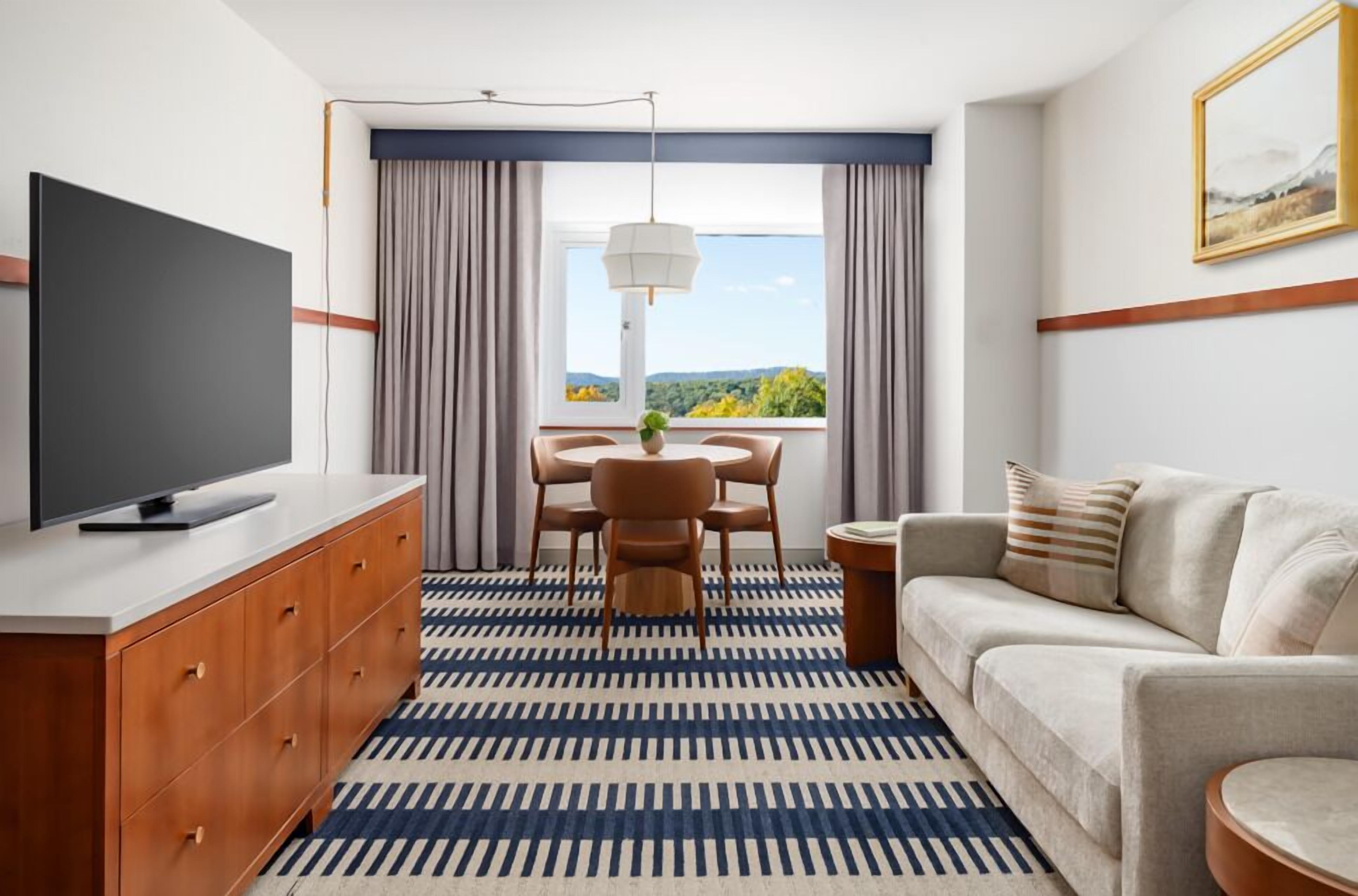 Ein modernes Hotelzimmer mit Fernseher, einer Couch, einem Esstisch mit Stühlen und einem großen Fenster mit Vorhängen.