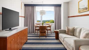 Una habitación de hotel moderna con televisión de pantalla plana, un sofá, una mesa de comedor con sillas y un ventanal con cortinas.