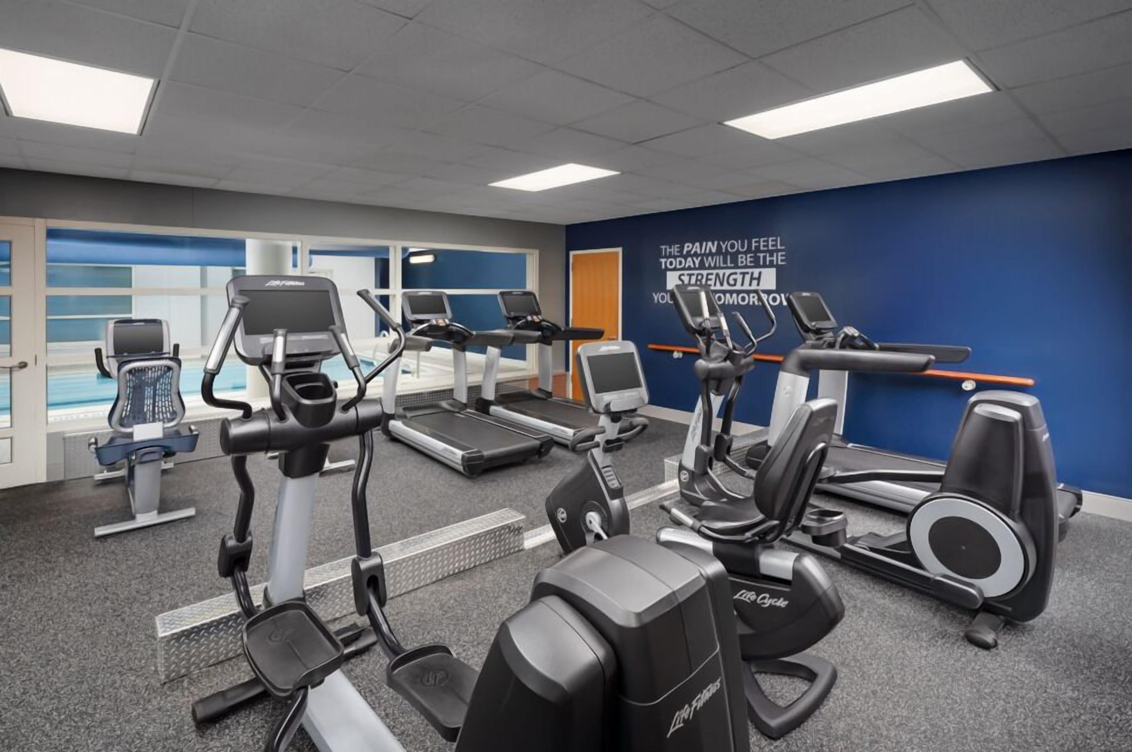 A gym with various cardio machines, a motivational quote on the wall, and a view of a pool through the windows.