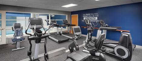 Une salle de sport équipée de divers appareils de cardio, une citation motivante affichée sur le mur et une vue sur une piscine par les fenêtres.