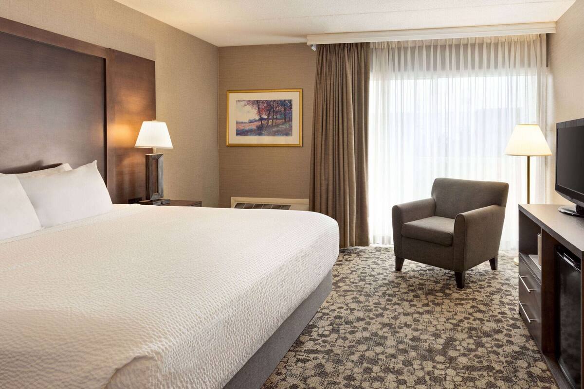 A hotel room with a large bed, a chair, a desk with a TV, and a painting on the wall.