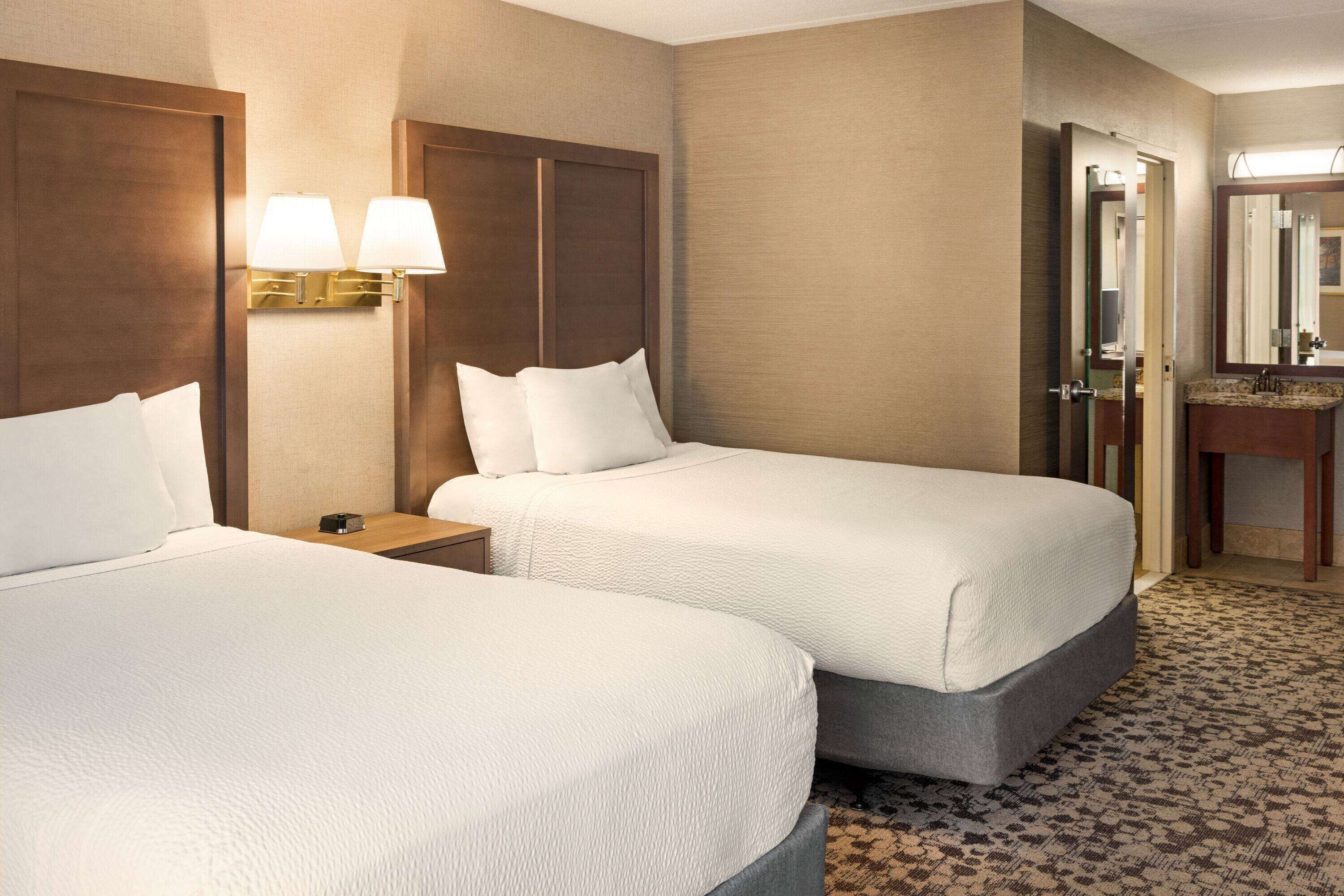 A hotel room with two beds, a nightstand, a lamp, a bathroom with a mirror, and a carpeted floor.