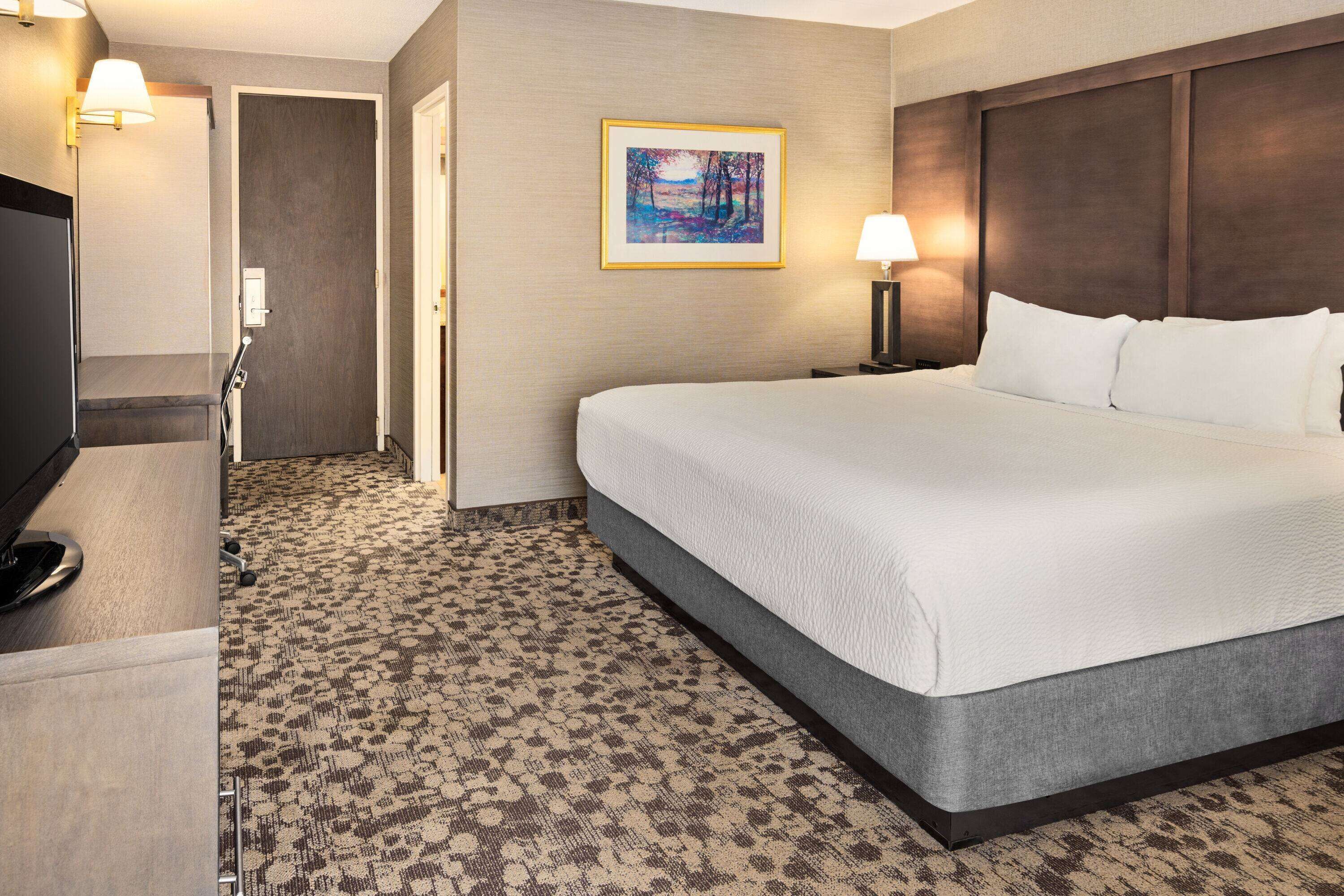 A hotel room with a large bed, a desk with a lamp, a painting on the wall, and a patterned carpet.