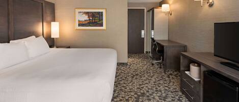 Habitación de hotel con una cama grande, un escritorio con televisor, una silla y un baño a la vista a través de una puerta abierta.