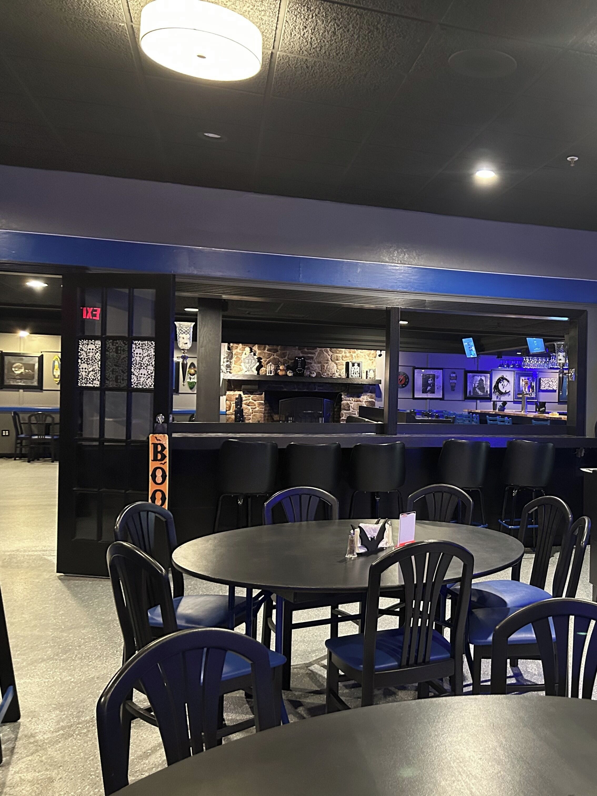 A restaurant with round tables and black chairs, a bar area with a stone wall, and an exit sign.